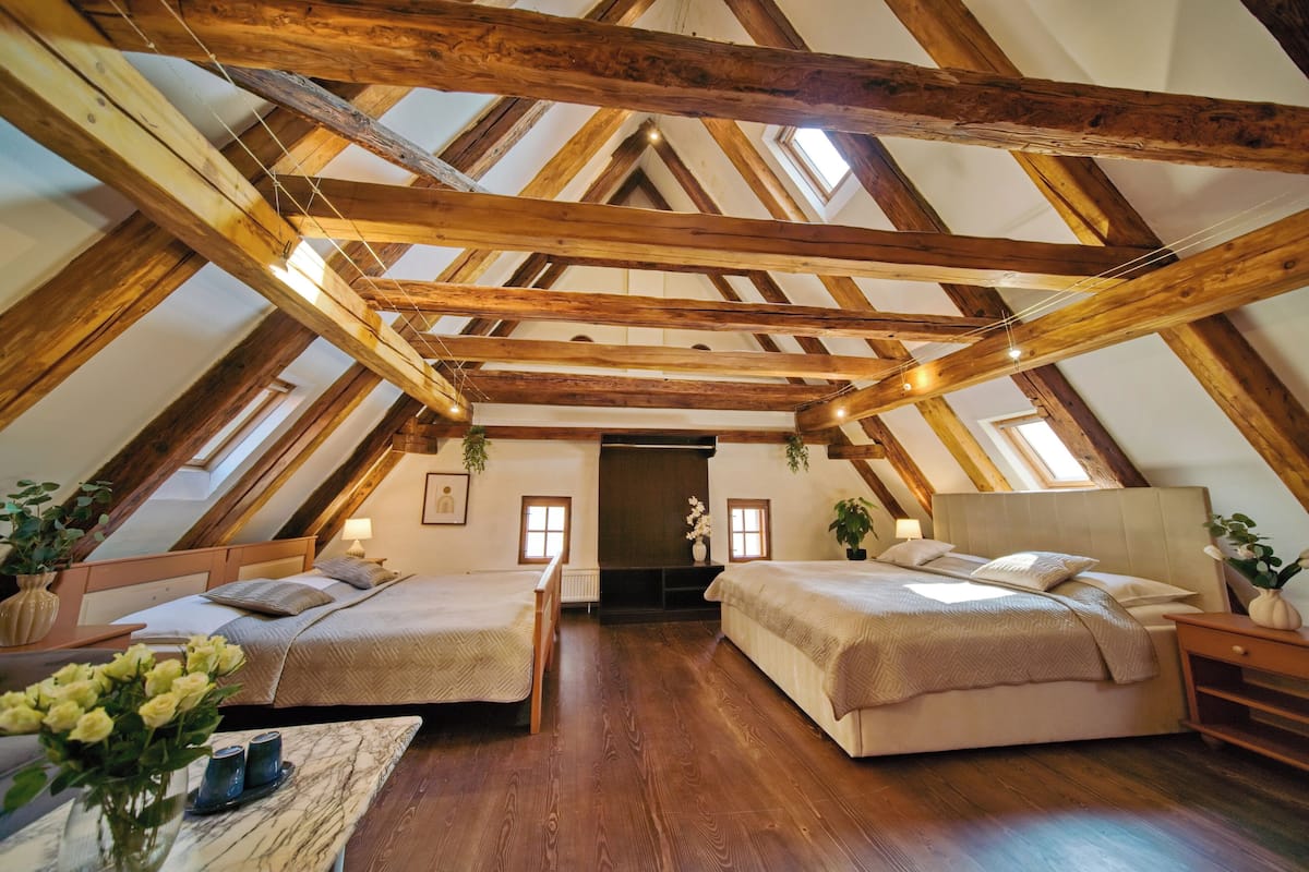 A high-ceilinged attic bedroom with exposed wooden beams, two beds, a marble table, and a view through a doorway.