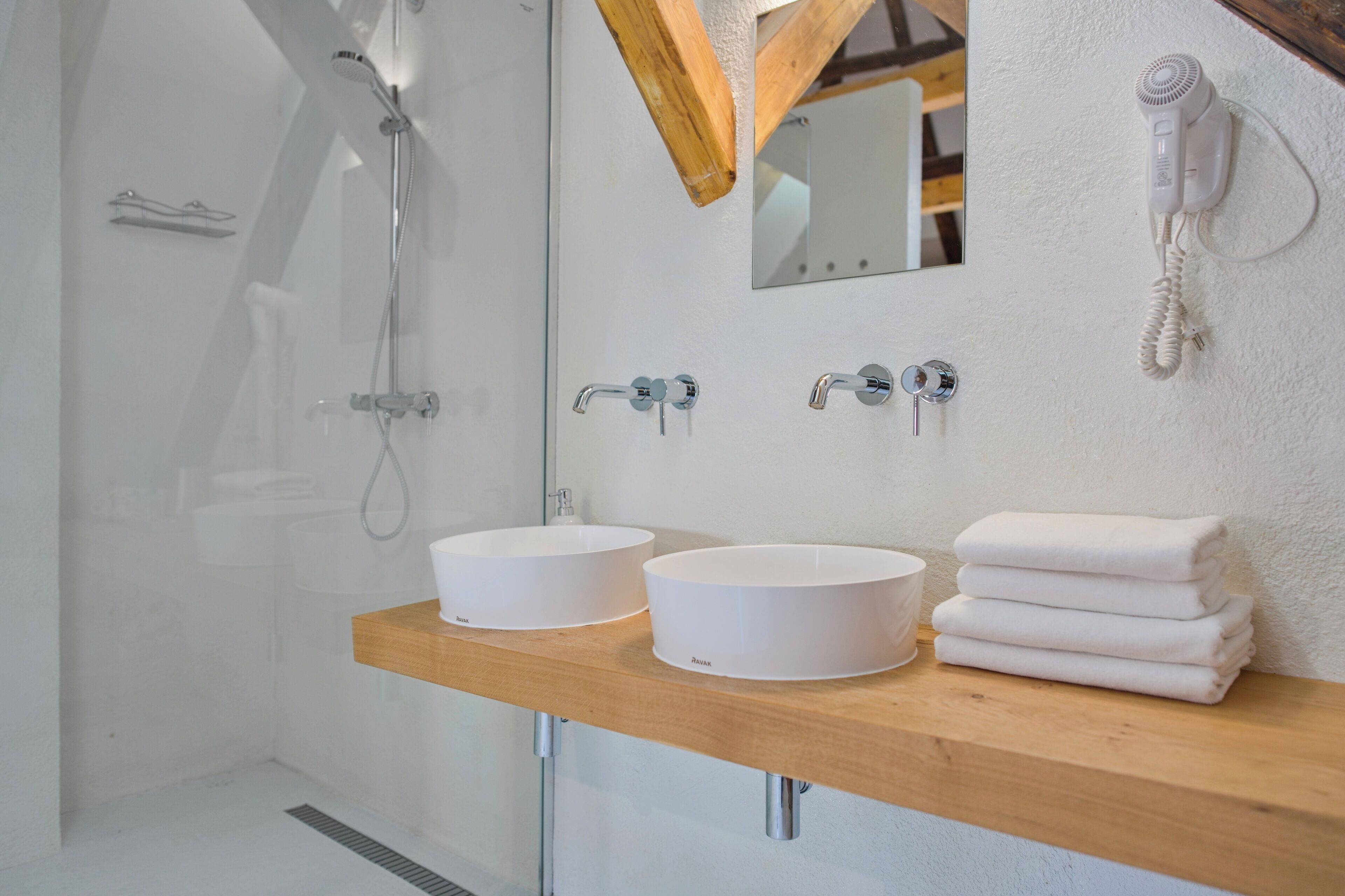 A modern bathroom with a glass-enclosed shower, two sinks, a wooden vanity, a mirror, and a hairdryer mounted on the wall.