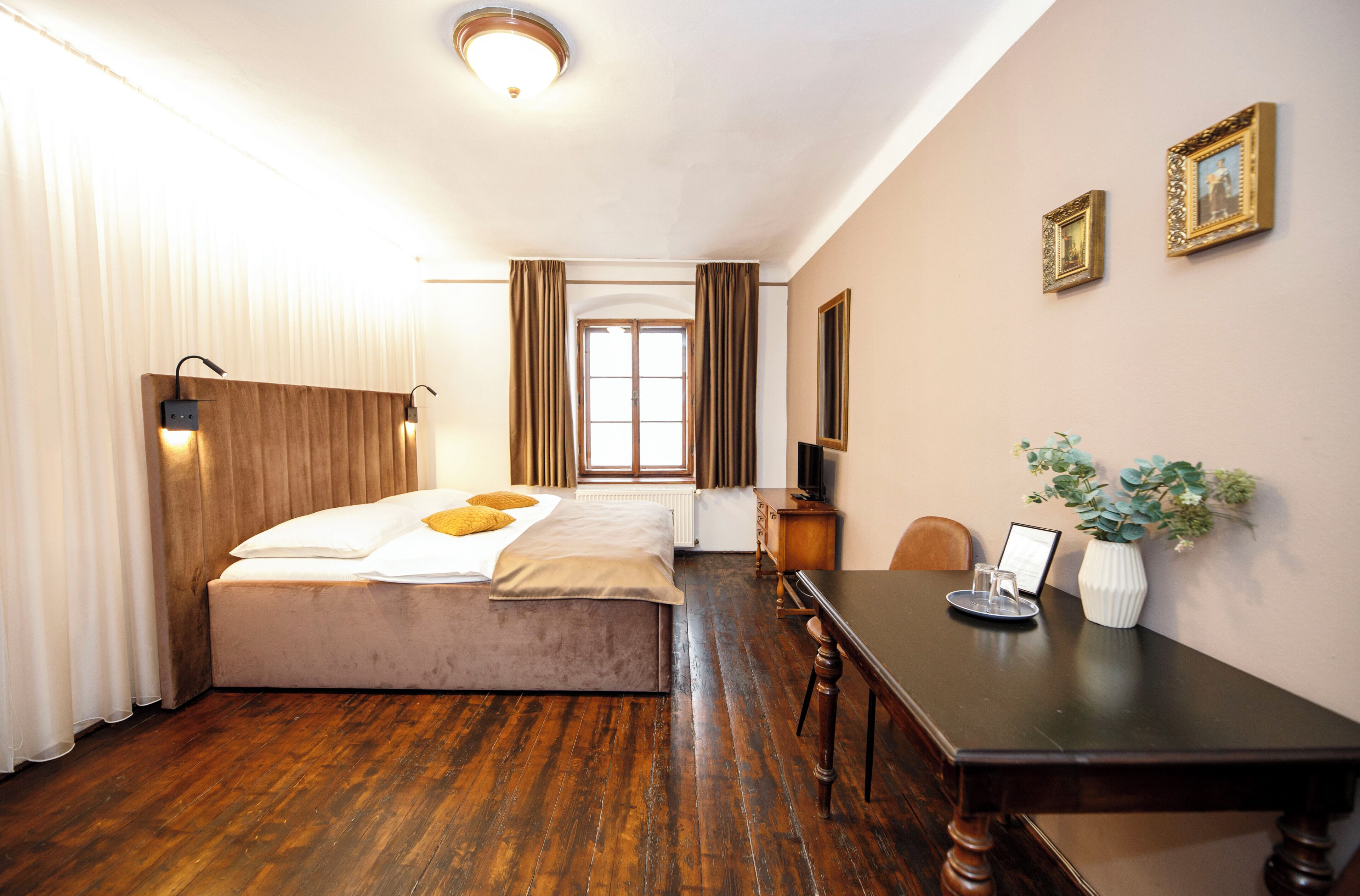 A hotel room with a bed, a wooden desk, a chair, a television, and a vase with plants.