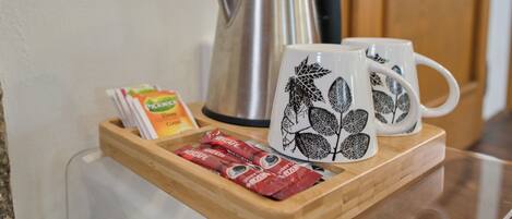 A tray with two white mugs with leaf designs, a stainless steel kettle, and packets of coffee and tea.