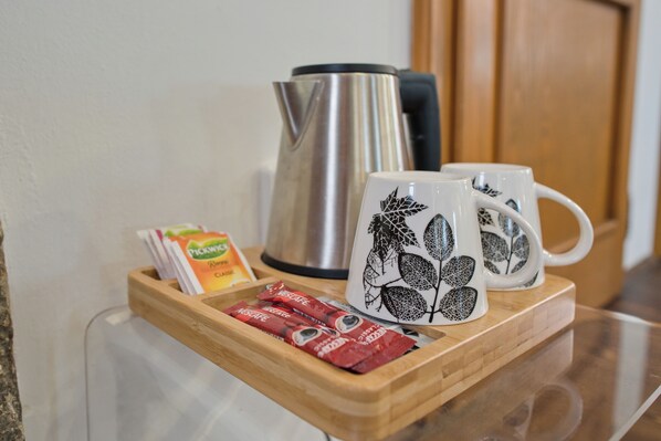 A tray with two white mugs with leaf designs, a stainless steel kettle, and packets of coffee and tea.