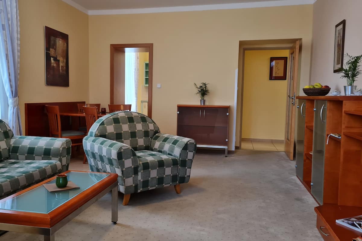 A living room with a sofa, armchair, and coffee table. There is a dining table with chairs, a cabinet, and a door leading to another room.
