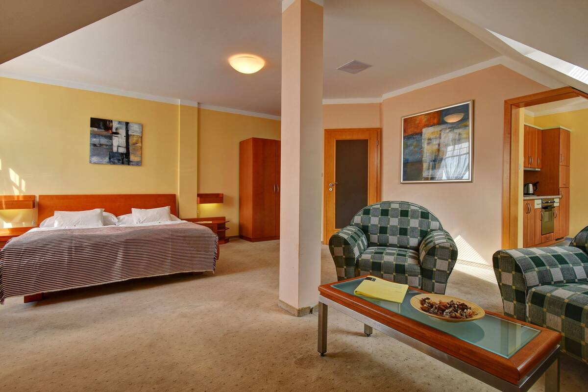 A hotel room with a bed, a checkered armchair, a small table with a plate of food, and a painting on the wall.