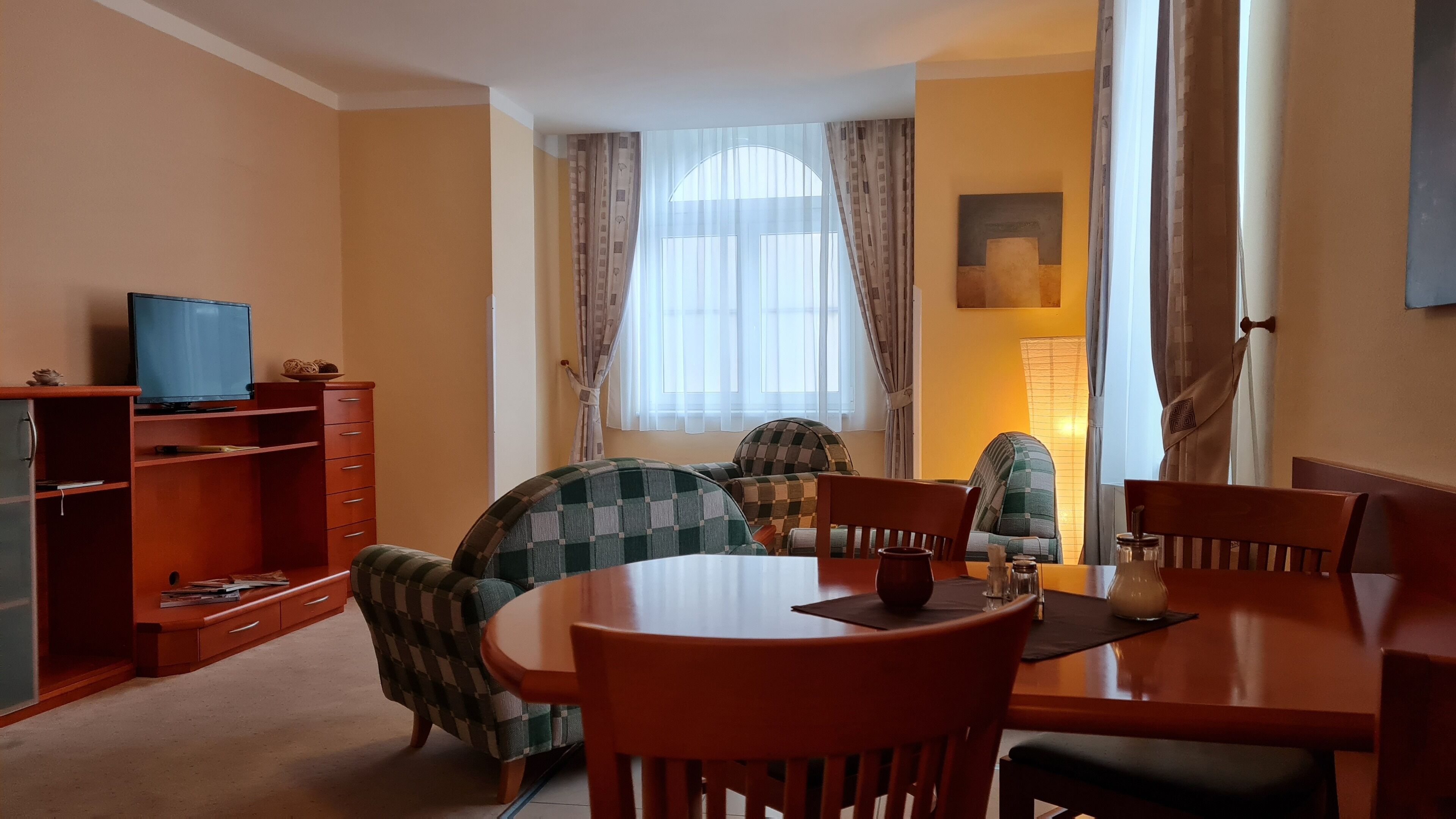 A dining area with a round table, four chairs, a television on a stand, and a window with curtains.