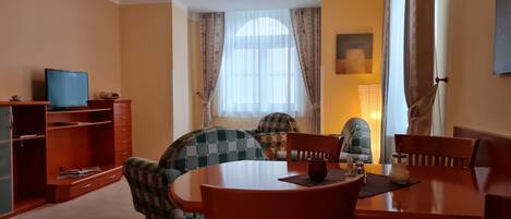 A dining area with a round table, four chairs, a television on a stand, and a window with curtains.