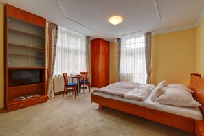 Un dormitorio con una cama, un televisor, un armario de madera, un escritorio con sillas y una ventana con cortinas.
