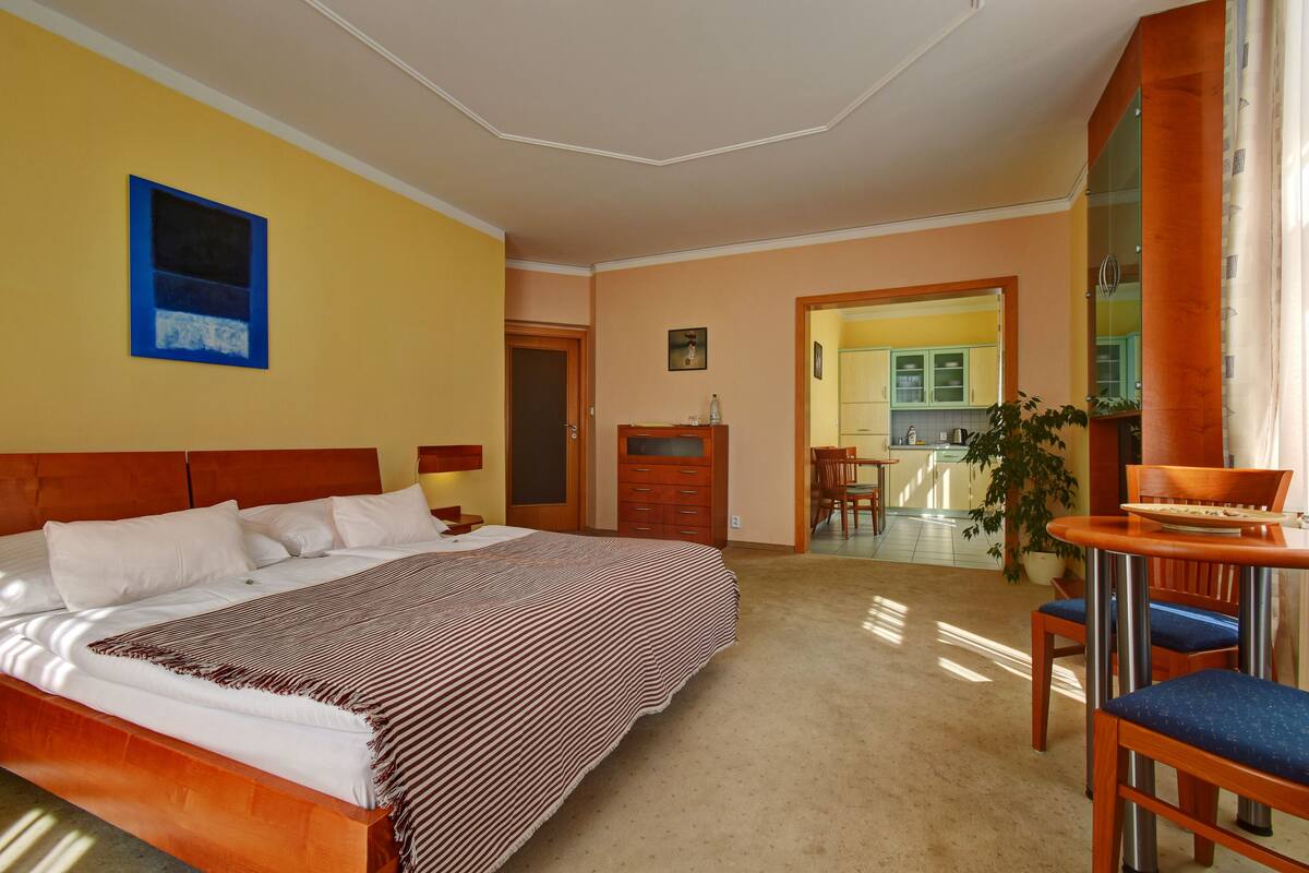 A hotel room with a large bed, a wooden headboard, a desk with a chair, and a view of the kitchen and dining area.