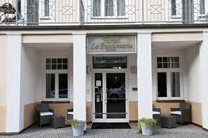 La entrada del Hotel La Passionaria, con un escaparate de vidrio, dos plantas en macetas y un coche estacionado afuera.