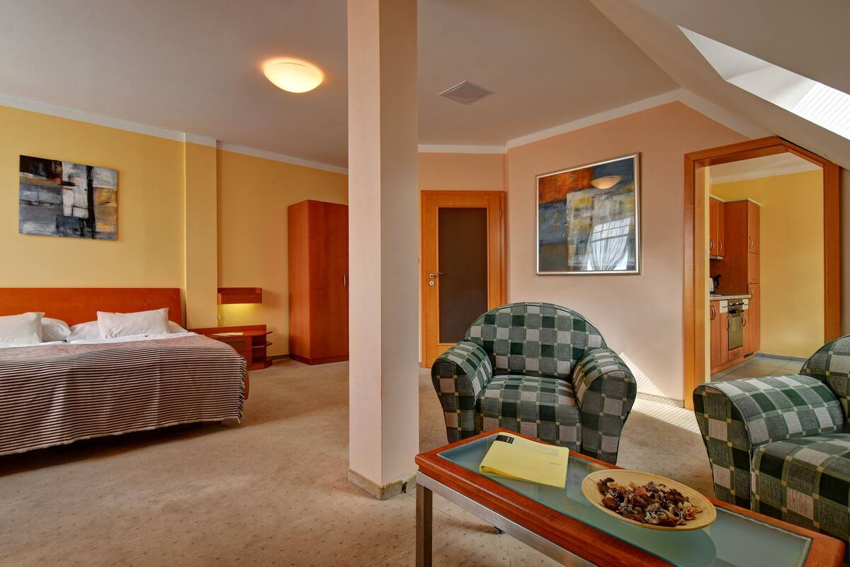 A hotel room with a bed, two checkered armchairs, a small table with a plate of food, a painting on the wall, and a door leading to a kitchen.