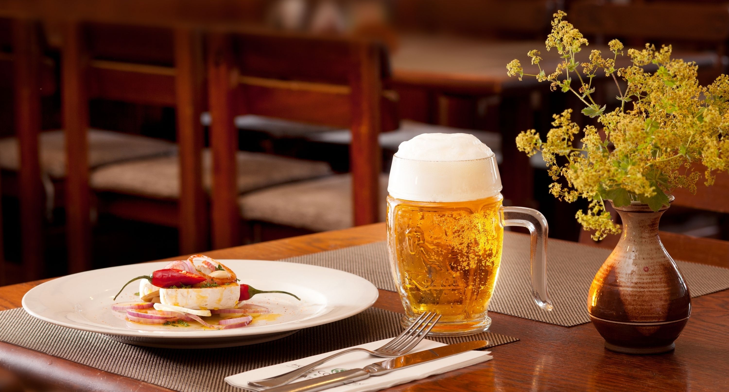Una mesa de comedor con un plato de comida, una jarra de cerveza y un florero con flores.