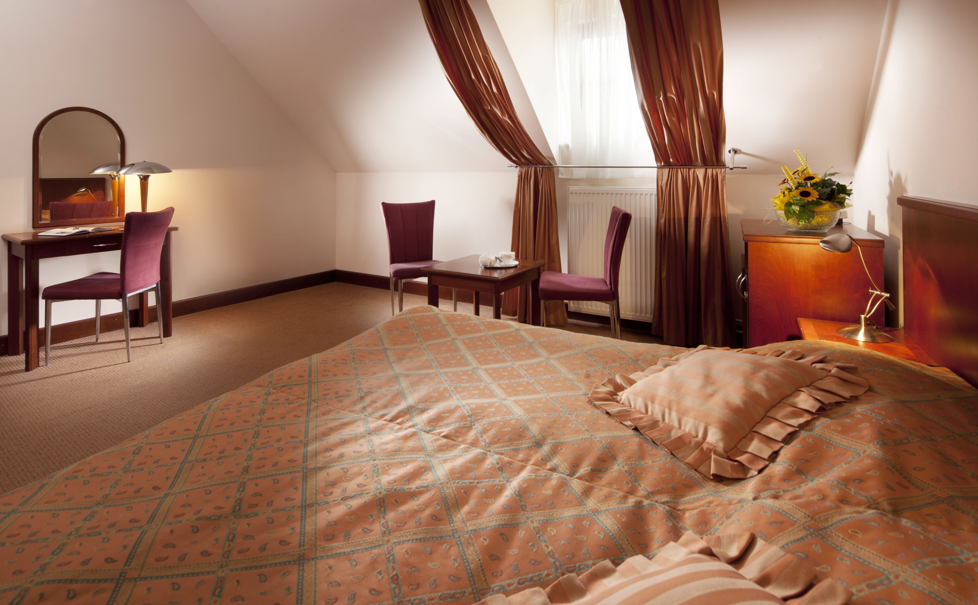 A hotel room with a large bed, a desk with a chair, a small table, and a lamp.
