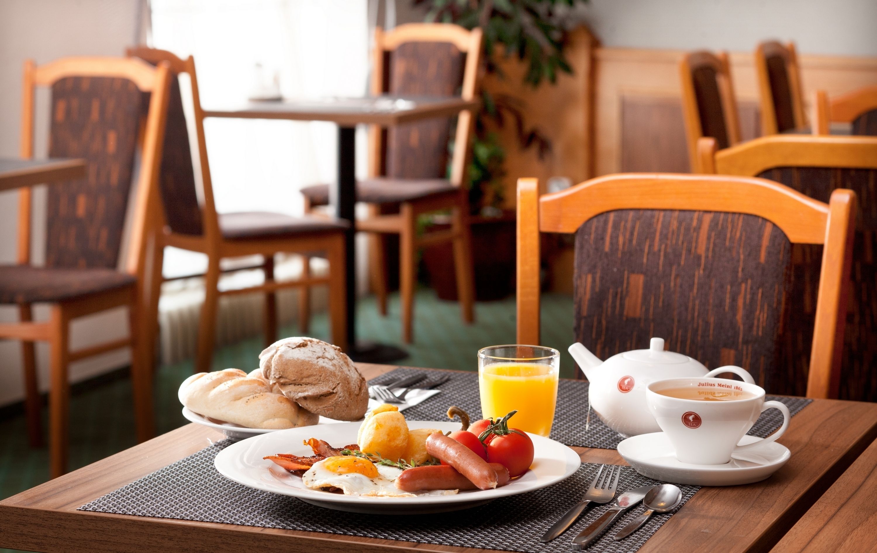 A breakfast setting with a plate of eggs, bacon, sausages, tomatoes, and bread rolls, a glass of orange juice, and a teapot with a cup of tea on a wooden table.