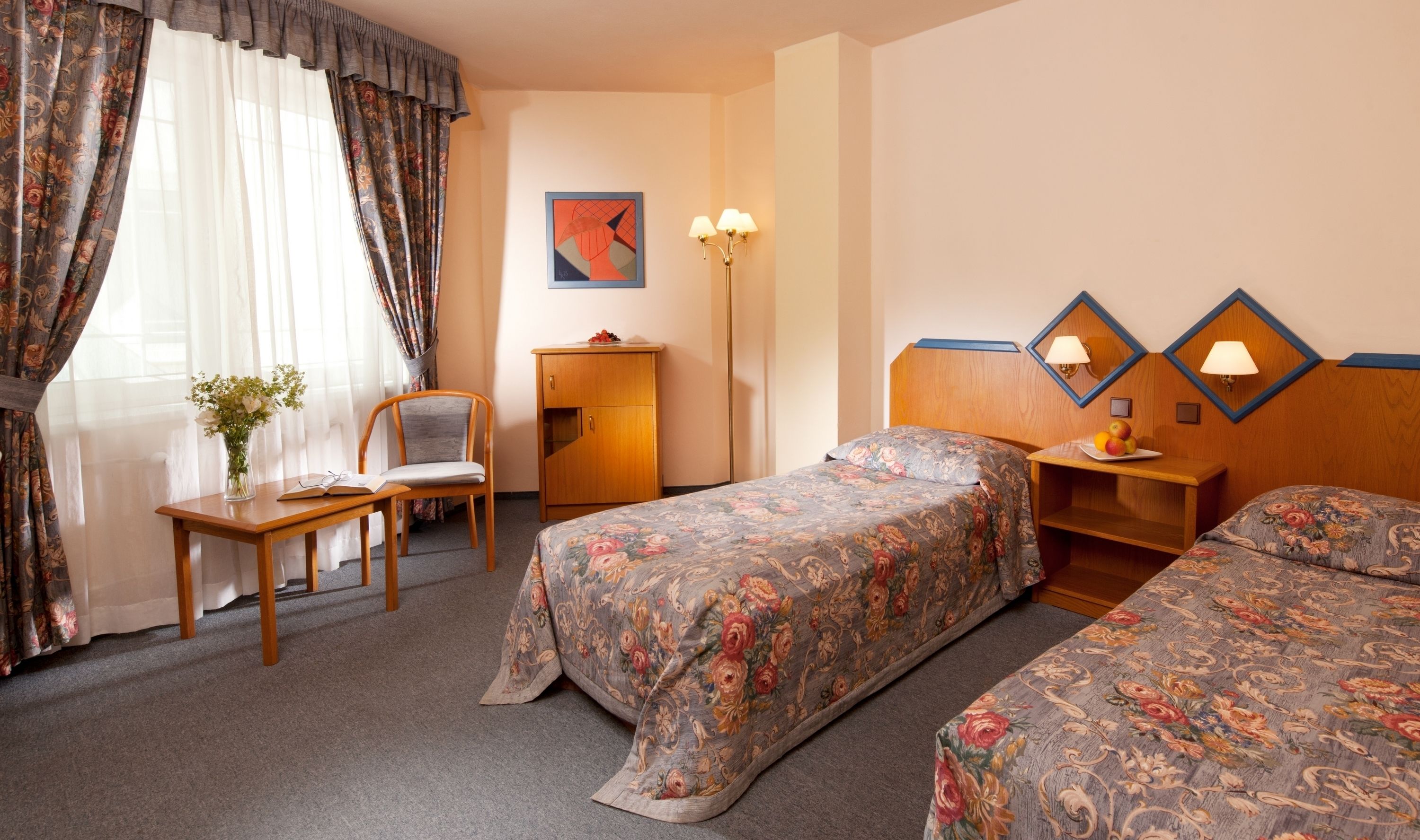 A hotel room with two beds, a desk, a chair, a small table with a vase of flowers, and a wall-mounted lamp.