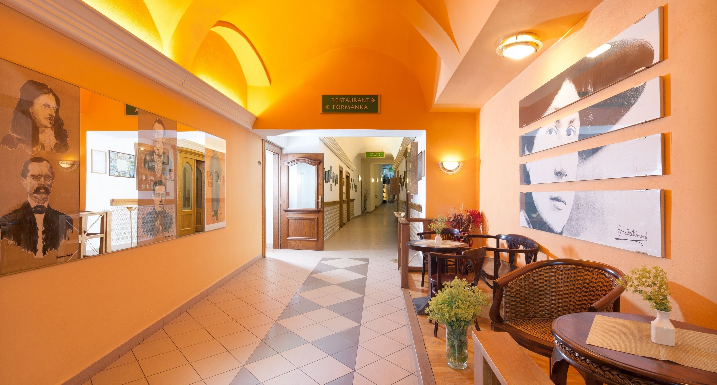 A hotel corridor with a checkered floor, framed portraits on the walls, a dining area with wooden furniture, and a sign indicating the direction to the restaurant.
