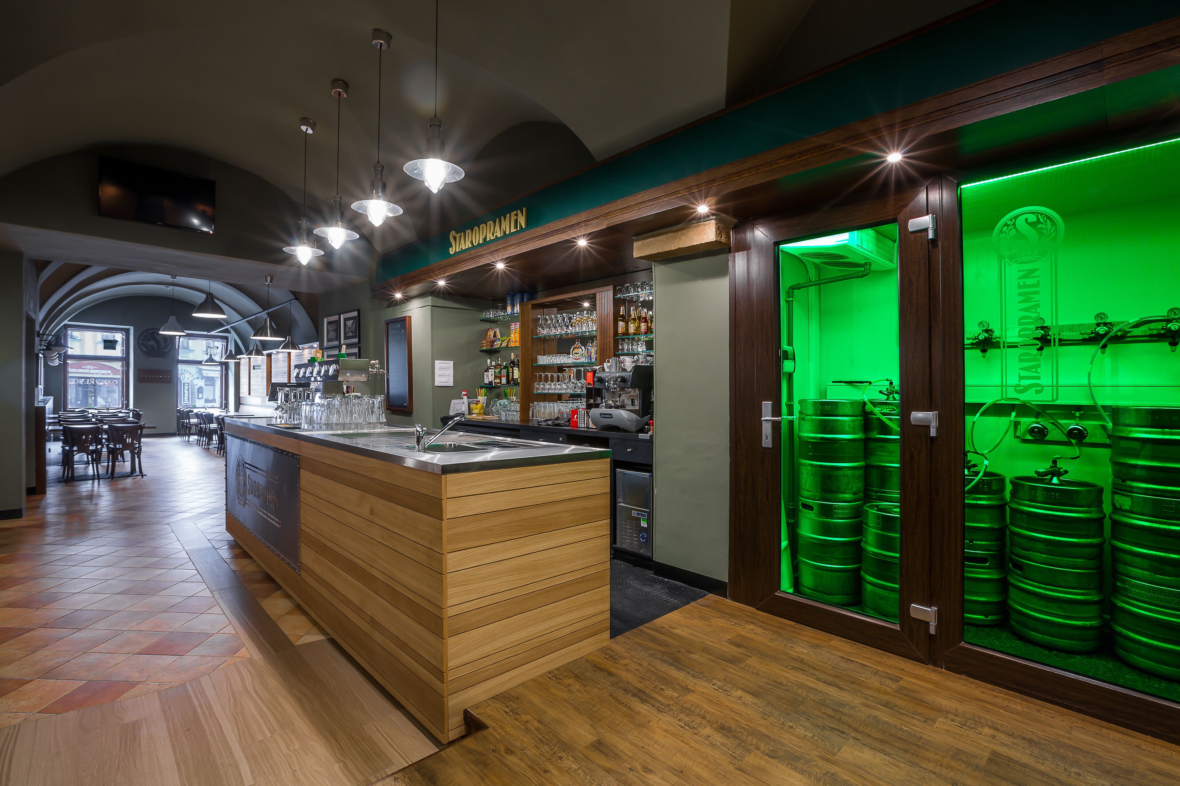 A bar area with a wooden counter, shelves with bottles, and a green-lit display of barrels.