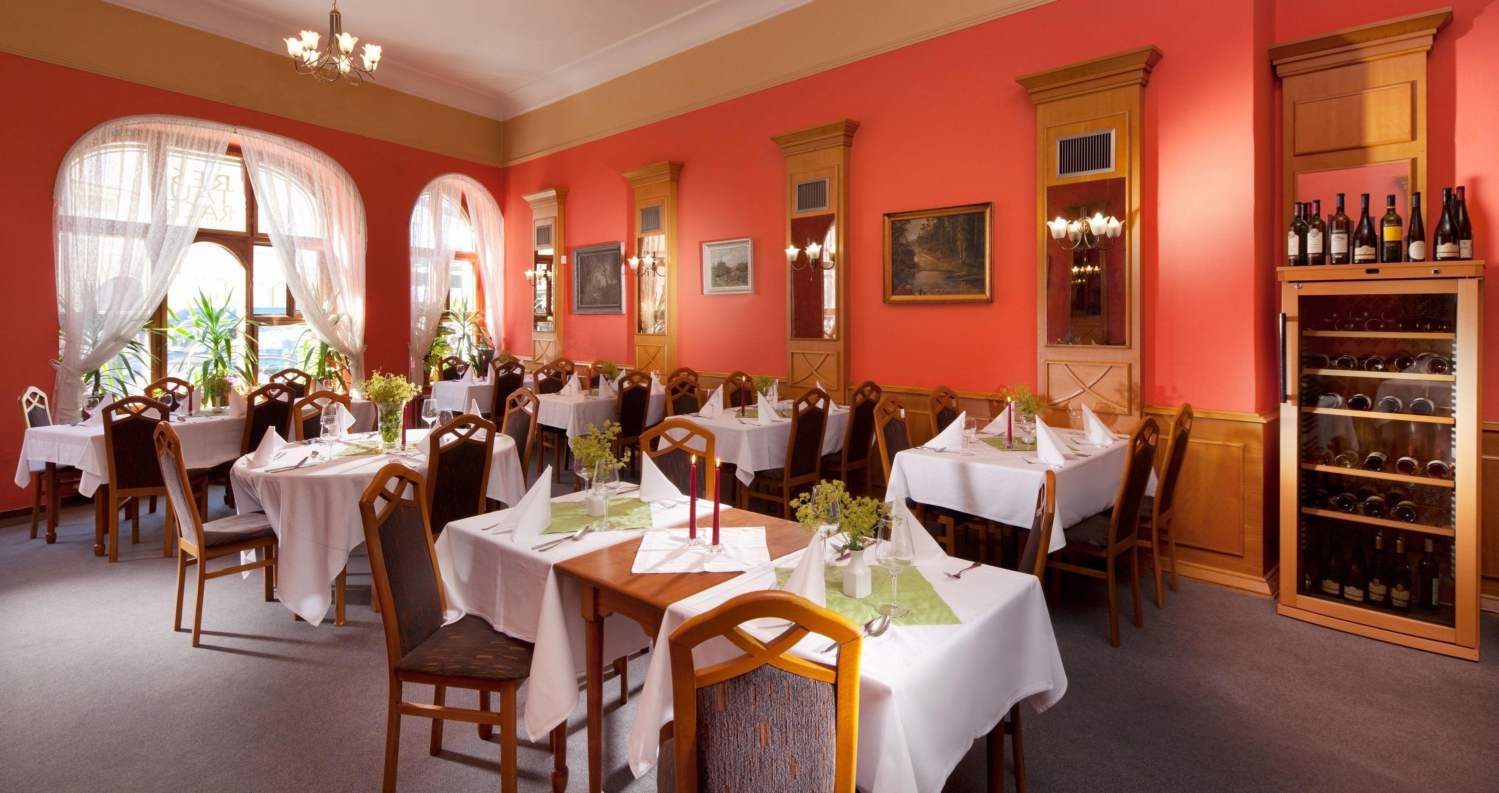 A dining area with multiple tables set for guests, featuring white tablecloths and wooden chairs.