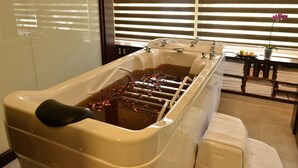 A spa bath with rose petals, a window with blinds, and a shelf with towels and toiletries.