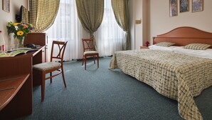 A hotel room with a bed, a desk, a chair, a desk lamp, and a vase with flowers.