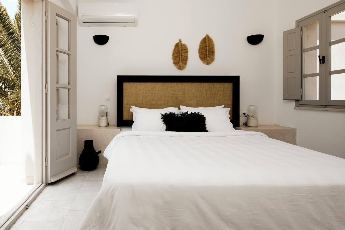 A bedroom with a bed, bedside tables, a wall-mounted air conditioner, and a window with open shutters.