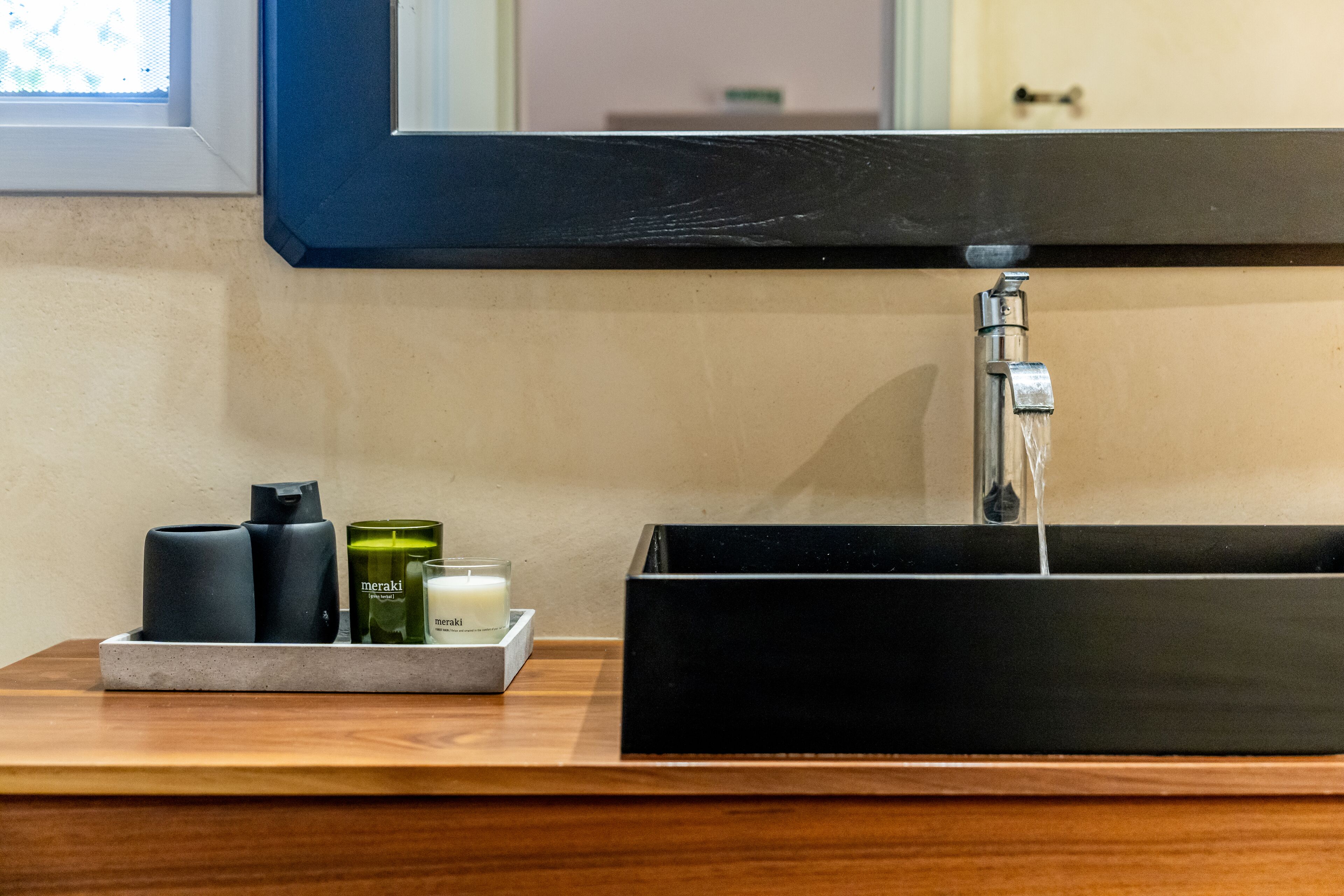 Un lavabo negro con un espejo encima, una encimera de madera y una bandeja con dos botellas negras, una verde y una vela blanca.