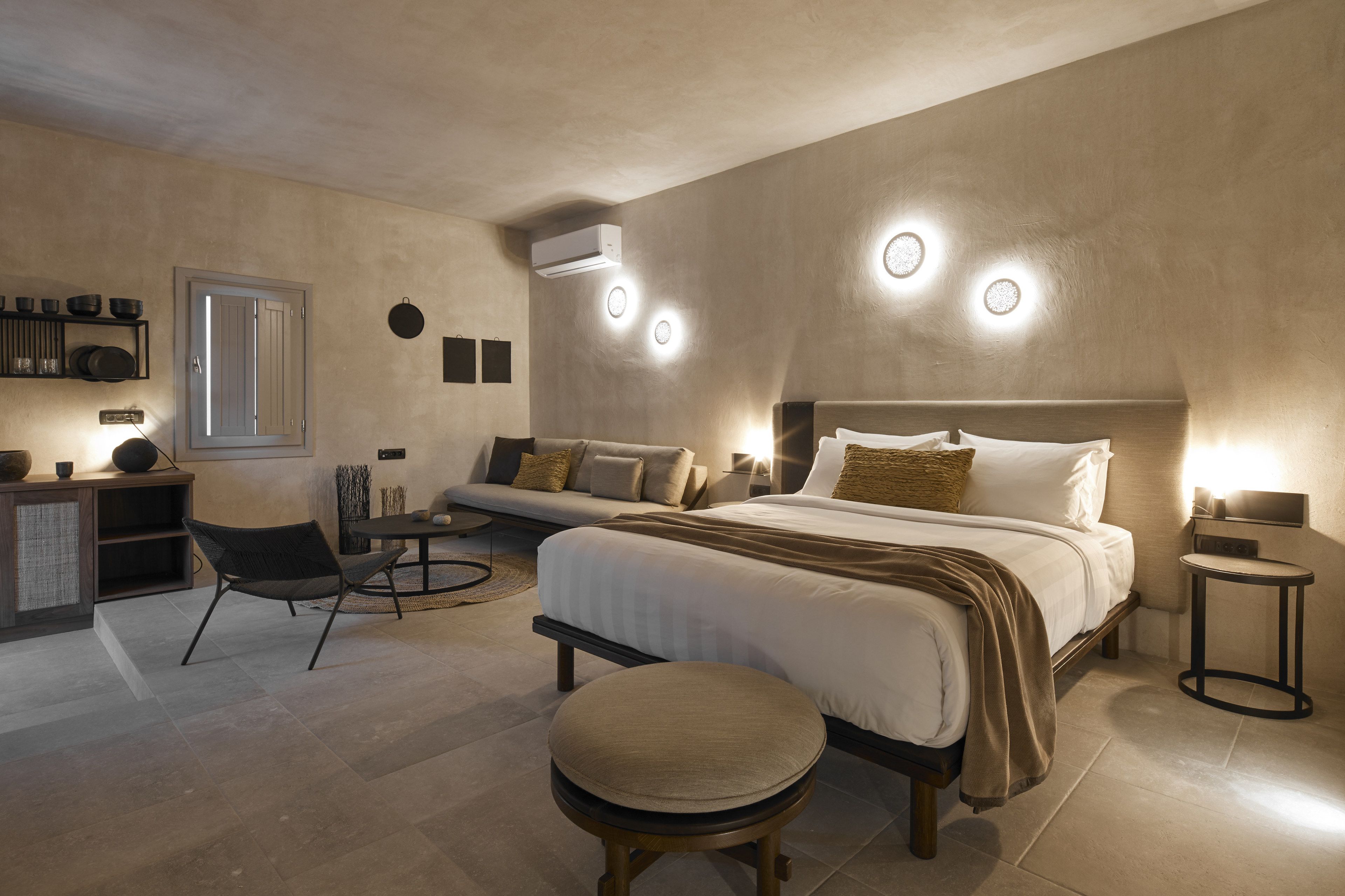 A modern bedroom with a large bed, a sofa, a dining table, and a small stool. There is a built-in shelf with kitchenware and a wall-mounted air conditioner.