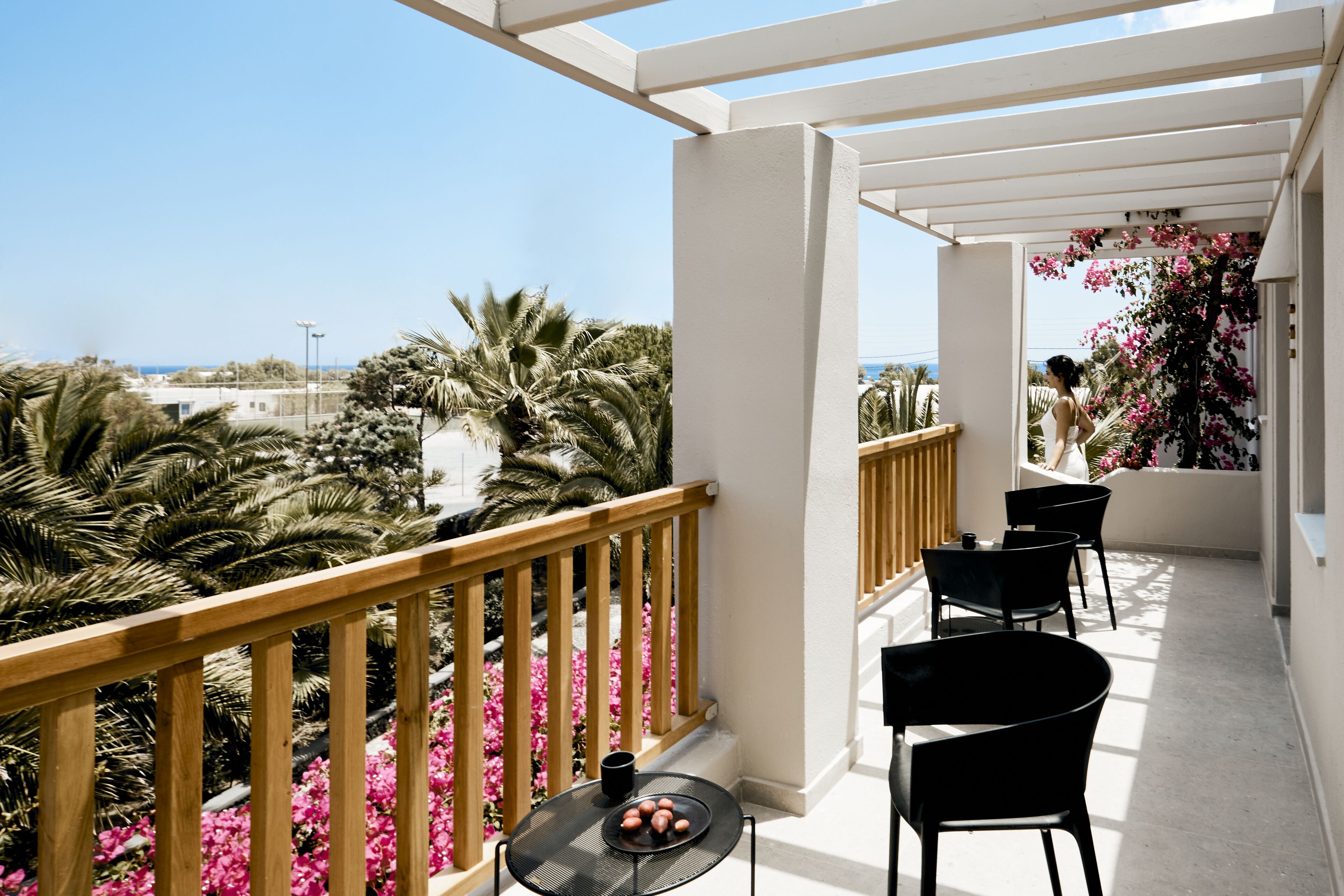 Ein Balkon mit Holzgeländer, einem kleinen Tisch mit Stühlen und Blick auf Palmen und Blumen.