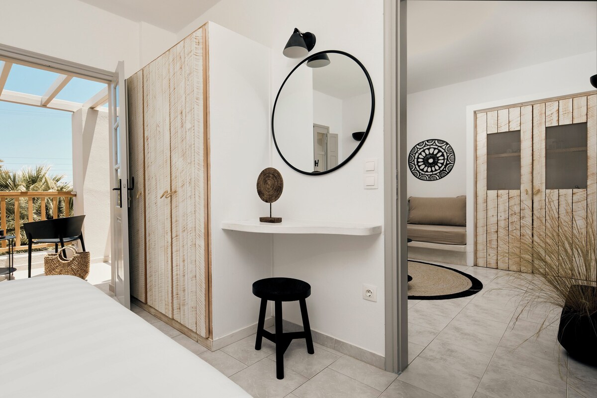 Un dormitorio moderno con una cama, un taburete negro, un espejo redondo, un estante con objetos decorativos y vistas al exterior.