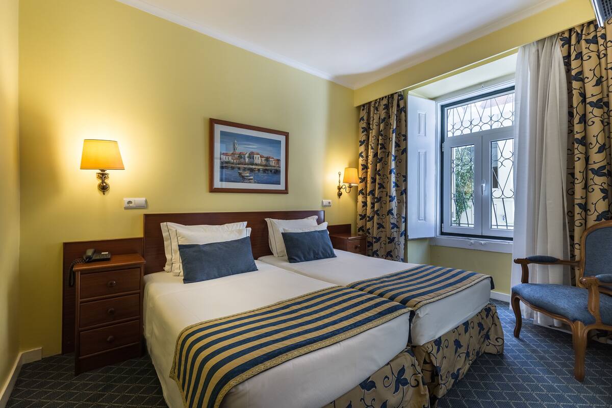 Habitación de hotel con una cama doble, dos mesitas de noche, una silla, una ventana con cortinas y un cuadro en la pared.