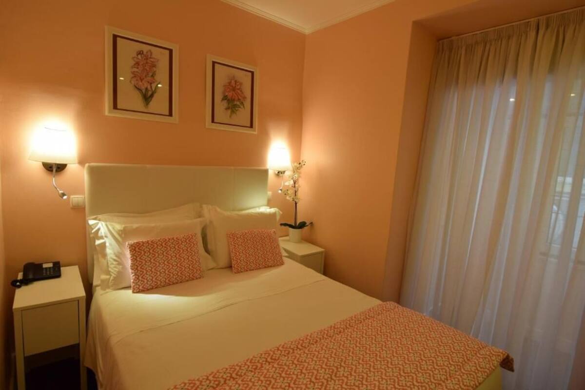 Una cama bien tendida con ropa de cama blanca y naranja, dos cuadros con motivos florales en la pared y una lámpara de noche a cada lado.