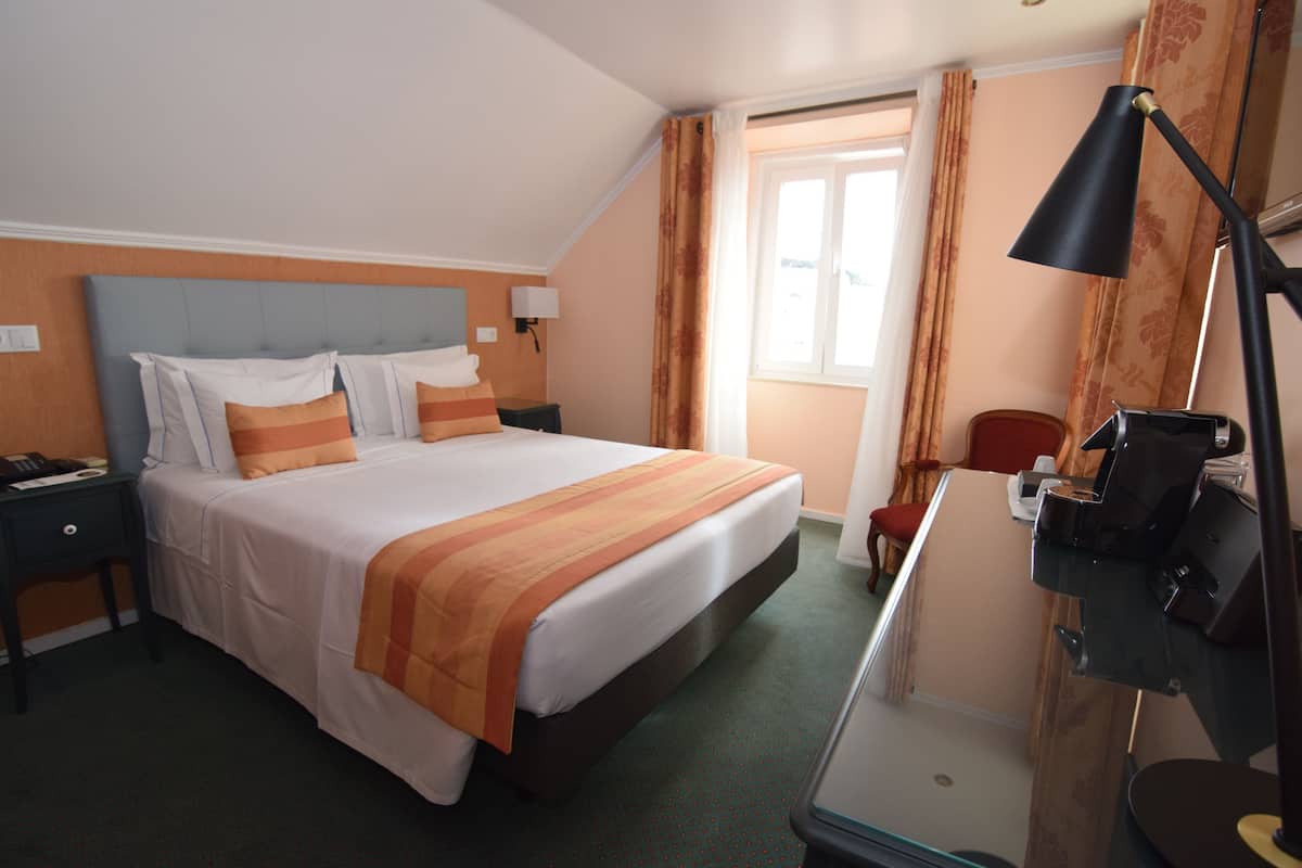 Habitación de hotel con una cama grande, un escritorio con lámpara, una silla y un ventanal con cortinas.