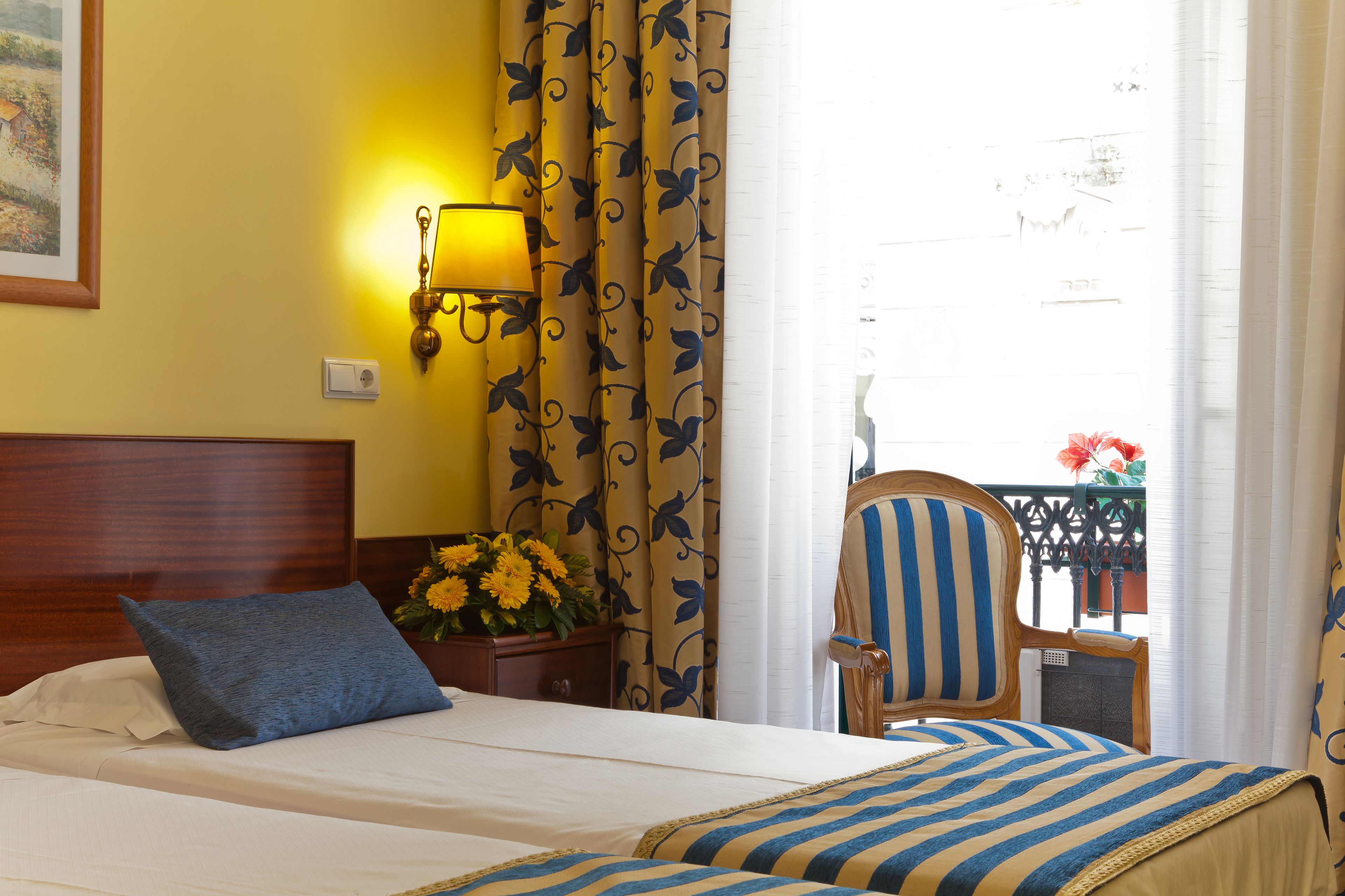 A hotel room with a bed, a nightstand with a lamp, curtains, and a view of the outside.