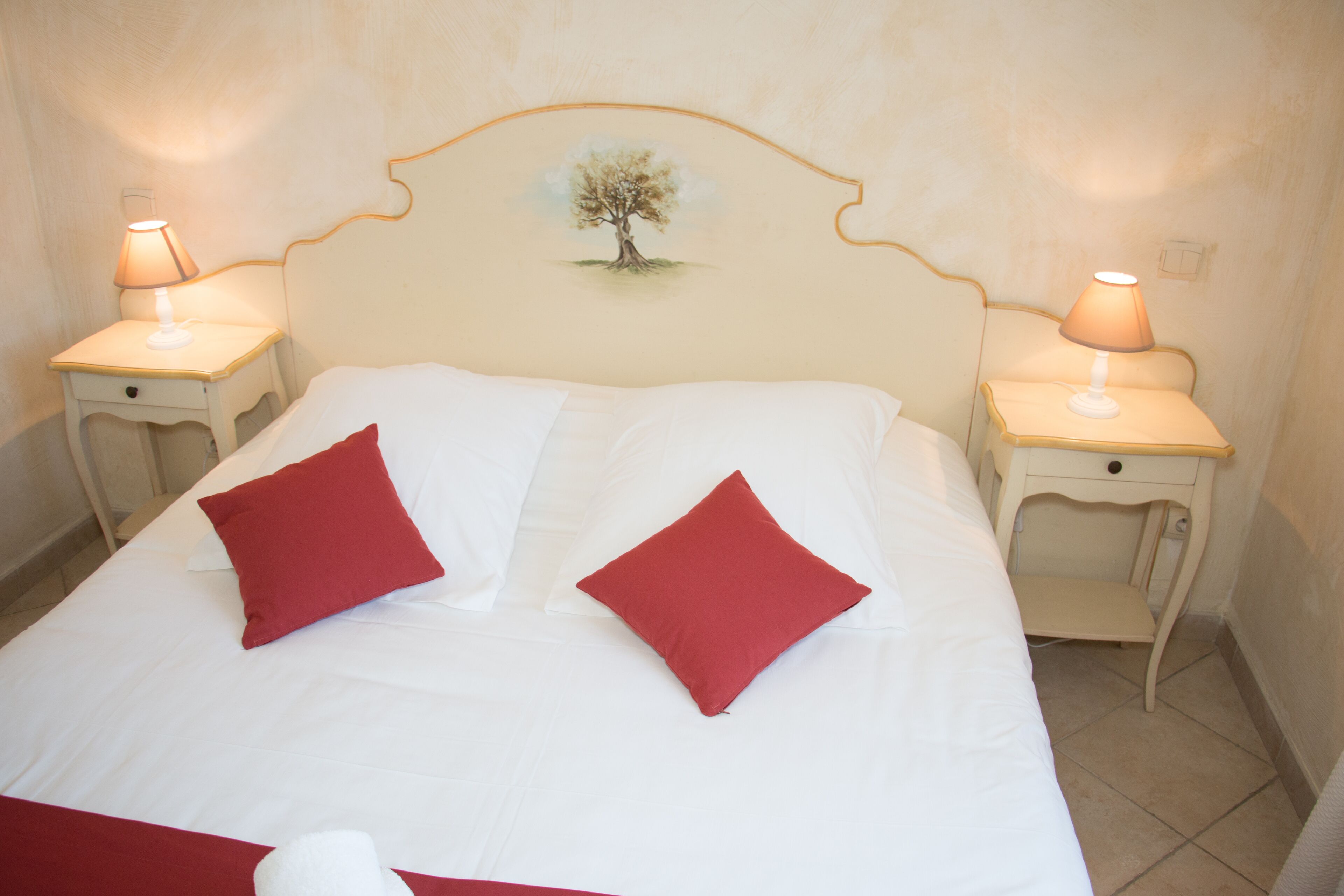 A neatly made bed with white and red pillows, two bedside lamps, and a wall-mounted artwork of a tree.