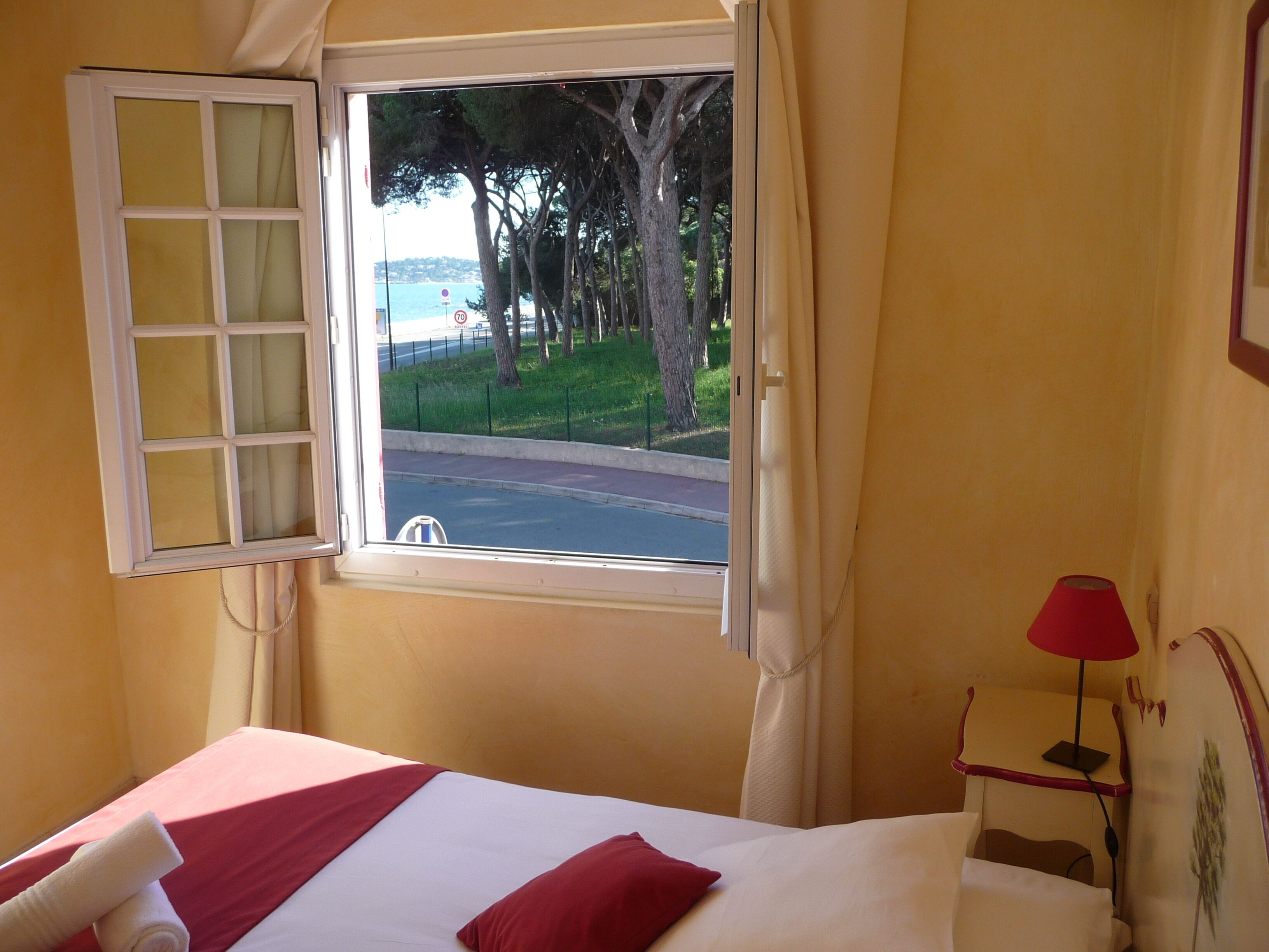 A hotel room with a bed, a window with curtains, a lamp, and a view of trees and a road.