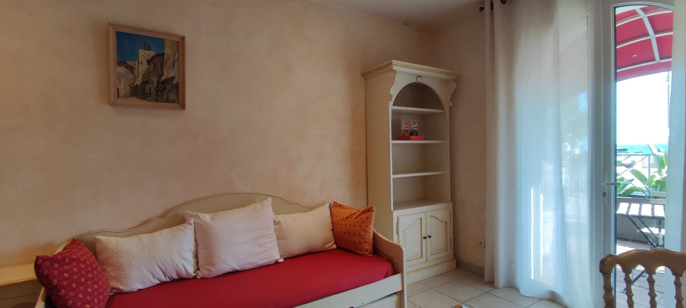 A room with a red sofa, a white cabinet, a painting on the wall, and a view through a window.