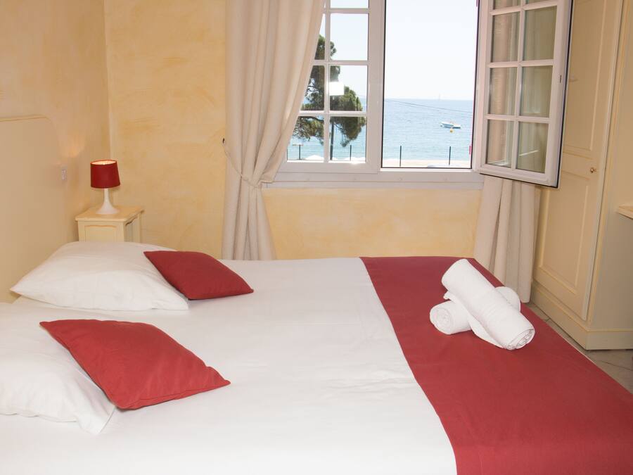A hotel room with a bed, a nightstand with a lamp, a window with a view of the sea, and a door.