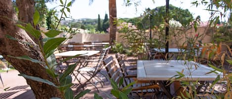 An outdoor patio with wooden tables and chairs, surrounded by trees and plants.