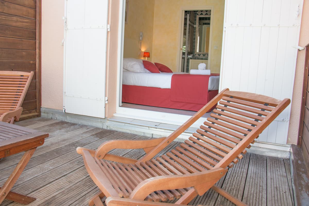 A wooden lounge chair on a wooden deck, a wooden table, and a room with a bed and a red blanket visible through an open door.