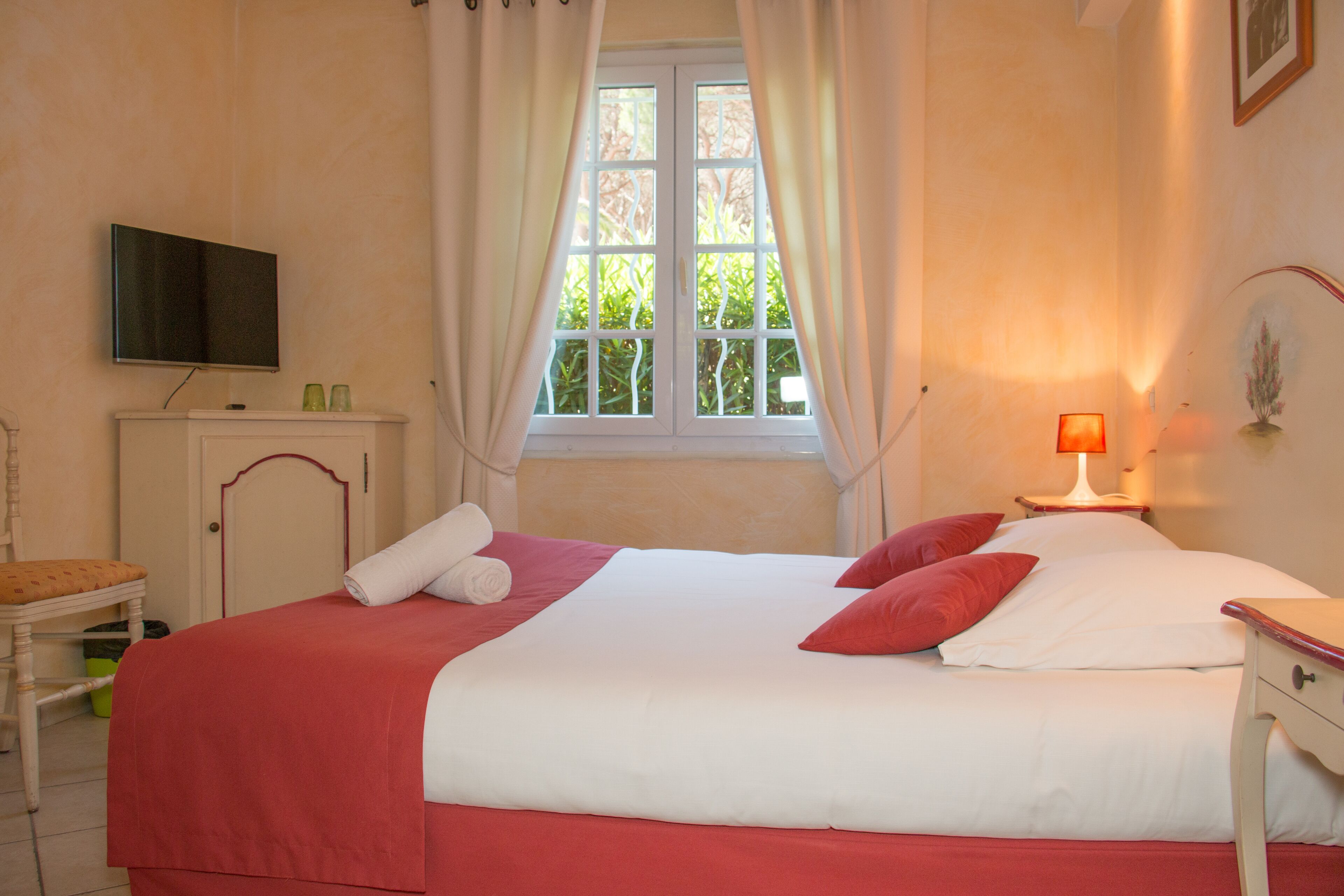 Una cama bien hecha con ropa de cama roja y blanca, una lámpara de noche, un televisor en la pared y una ventana con cortinas.