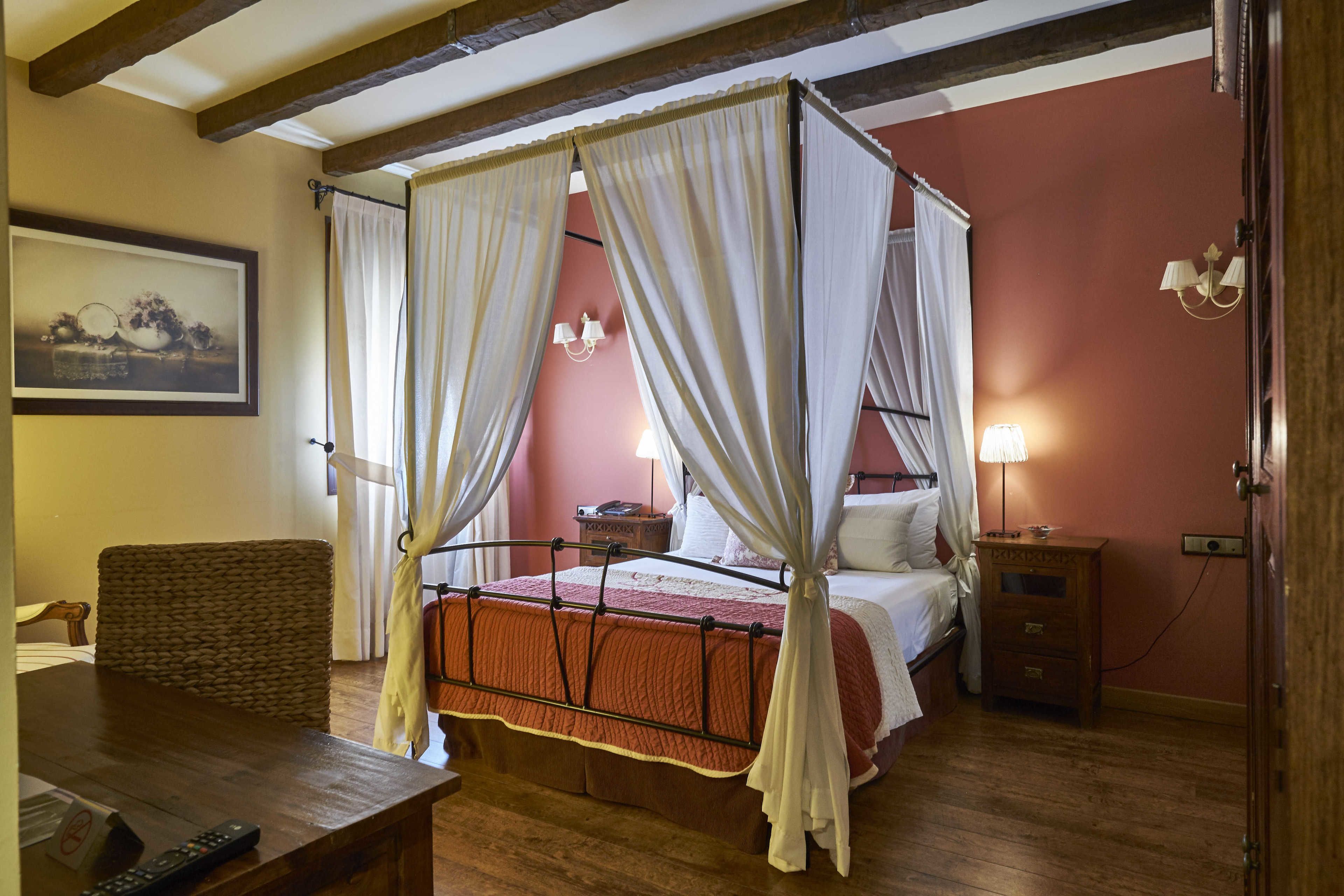 Een hemelbed met gordijnen, een houten nachtkastje met een lamp, een rieten stoel en een lijst met een afbeelding aan de muur.