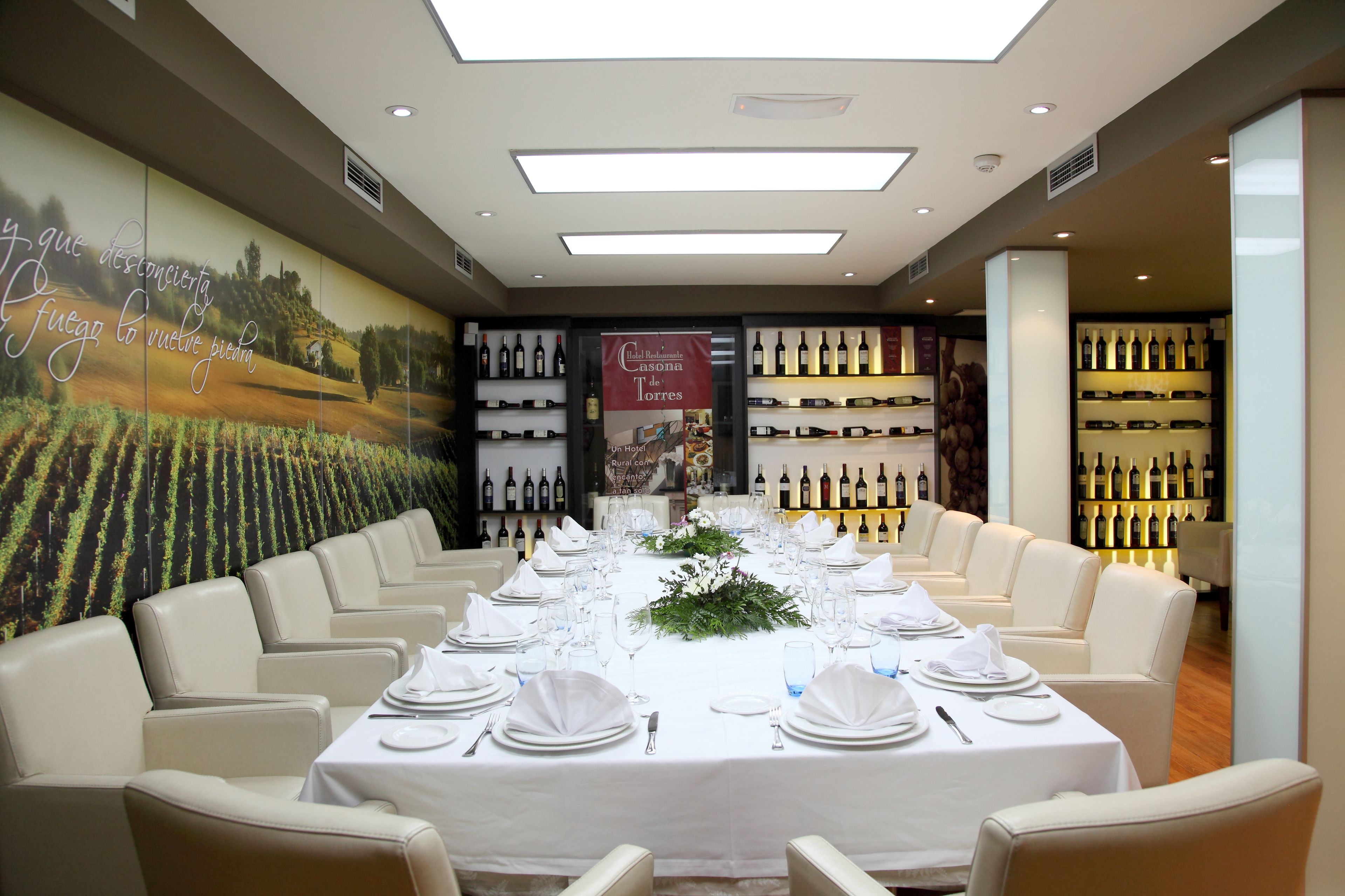 A dining area with a long table set for a meal, surrounded by chairs. There are wine bottles displayed on shelves in the background.