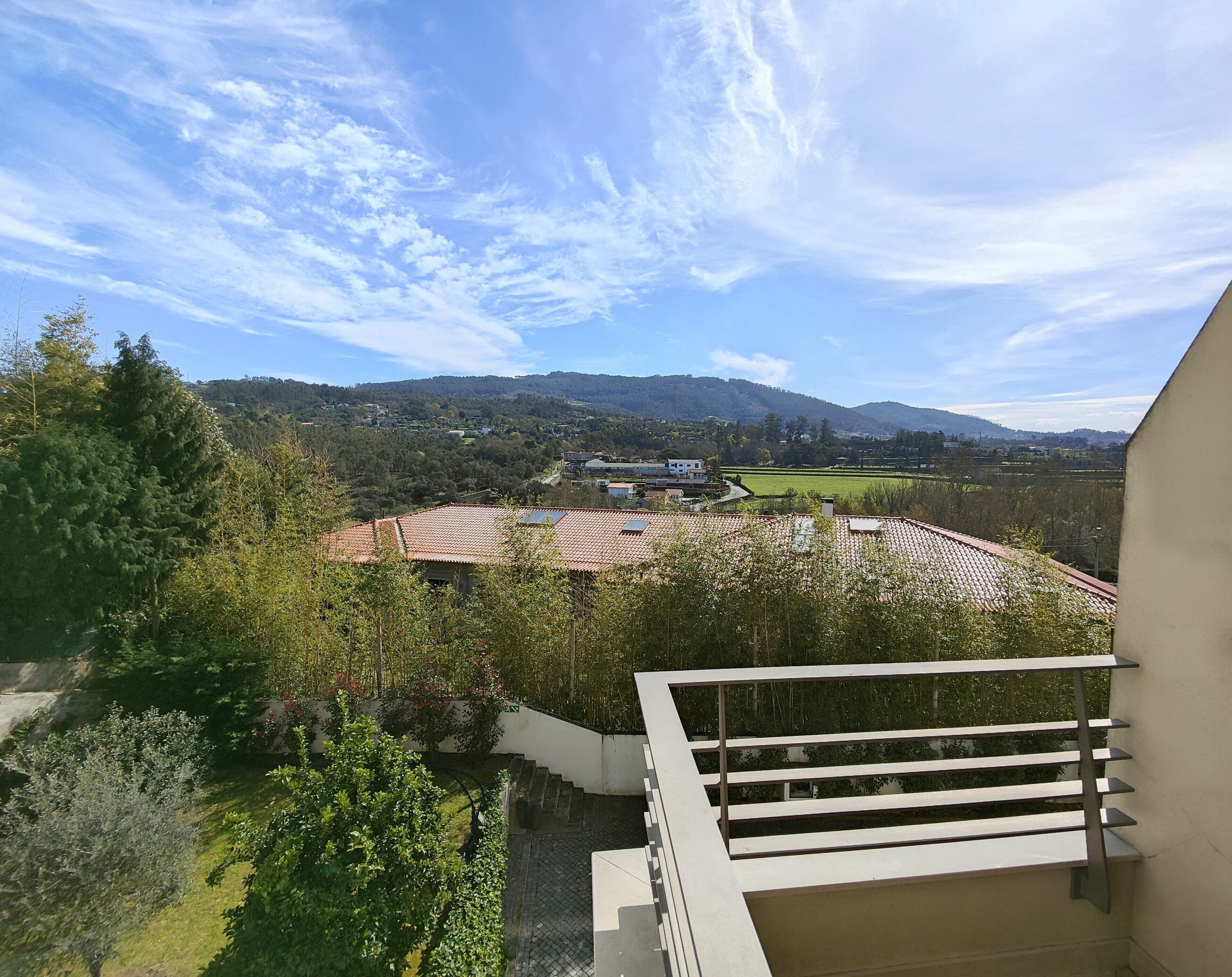 A balcony with a view of a green landscape, a building with a red roof, and a clear sky.