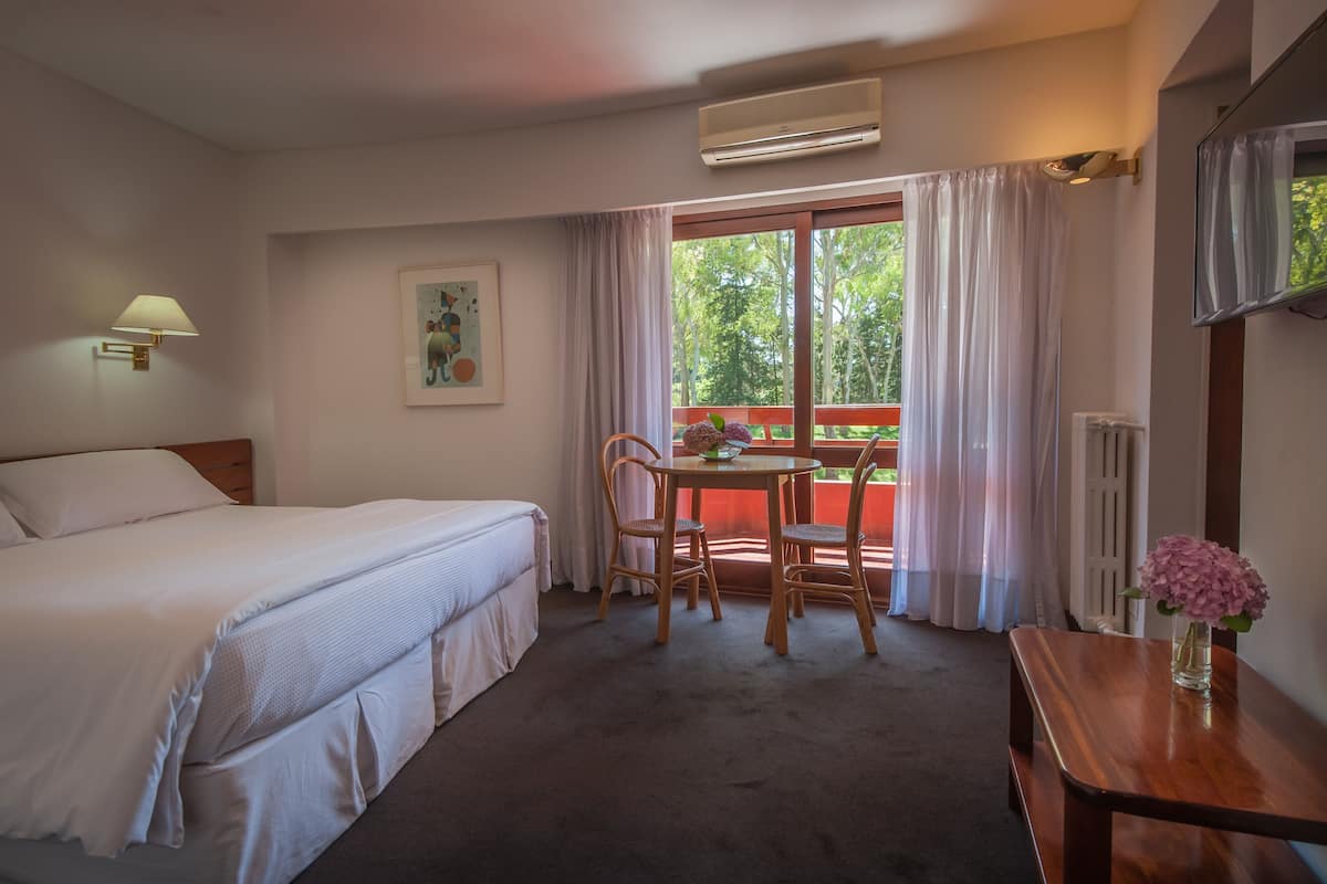 Una habitación de hotel con una cama, una mesa con sillas, una ventana con cortinas, un televisor montado en la pared y un radiador.