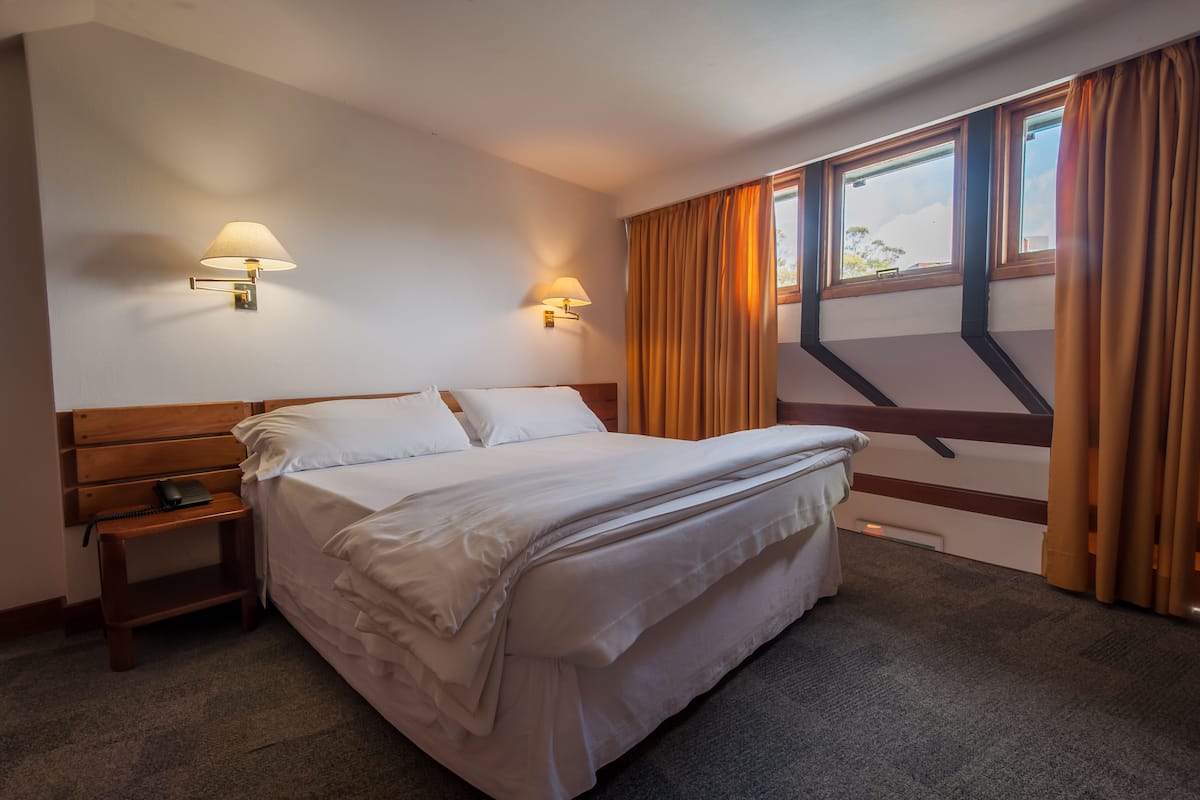 Habitación de hotel con una cama individual, dos lámparas fijadas a la pared, una mesita de noche con un teléfono y una ventana con cortinas.