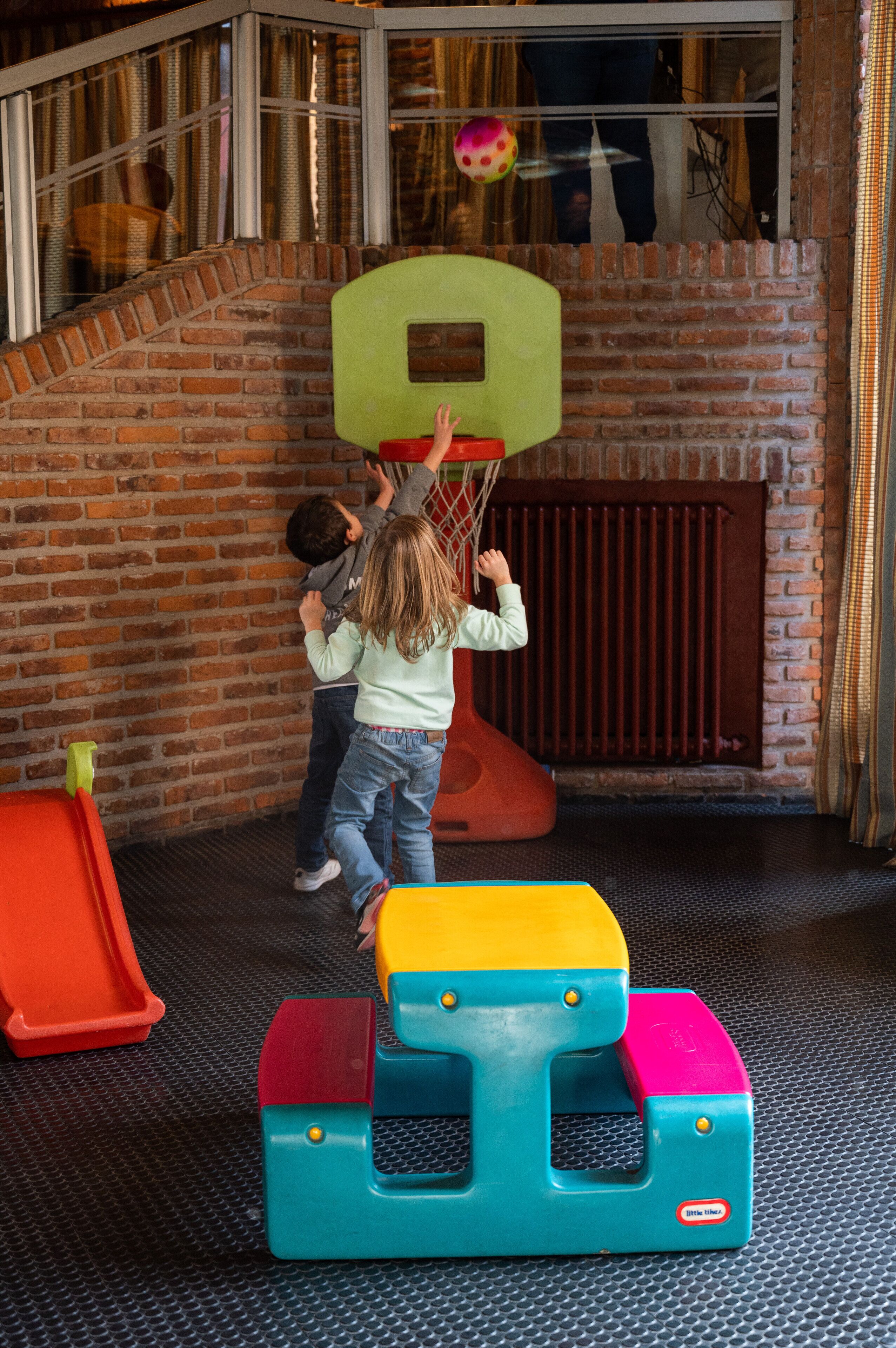 Des enfants jouent dans un parc, avec un mur de briques et une fenêtre en arrière-plan.