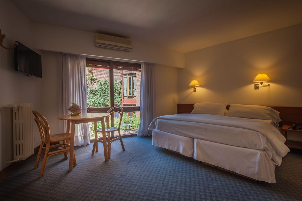 Habitación de hotel con una cama, dos lámparas de pared, una mesa con una silla, un televisor y un ventanal con cortinas.