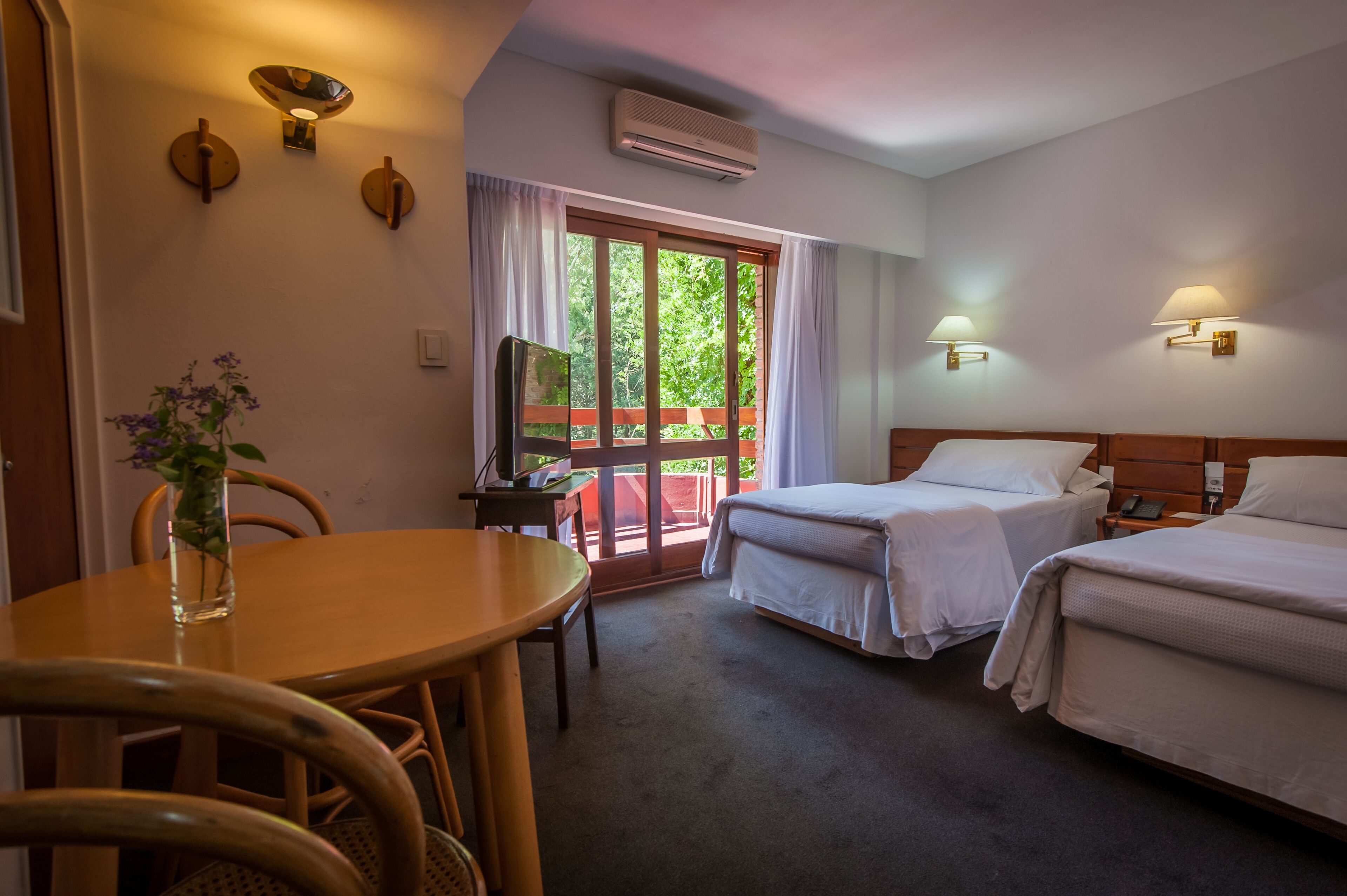 A hotel room with two beds, a dining table, chairs, a vase with flowers, and a view of greenery through the window.
