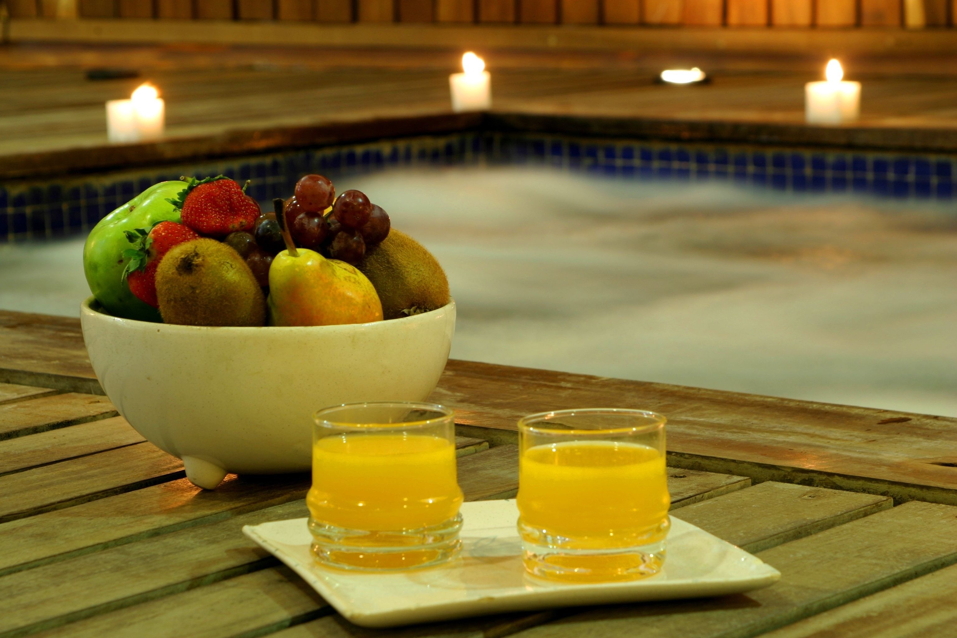 Un bol de fruta y dos vasos de jugo de naranja sobre una mesa de madera cerca de una piscina con velas.
