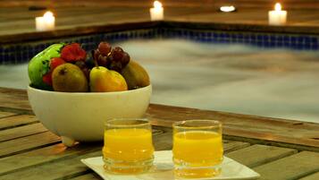 Un bol de fruta y dos vasos de jugo de naranja sobre una mesa de madera cerca de una piscina con velas.