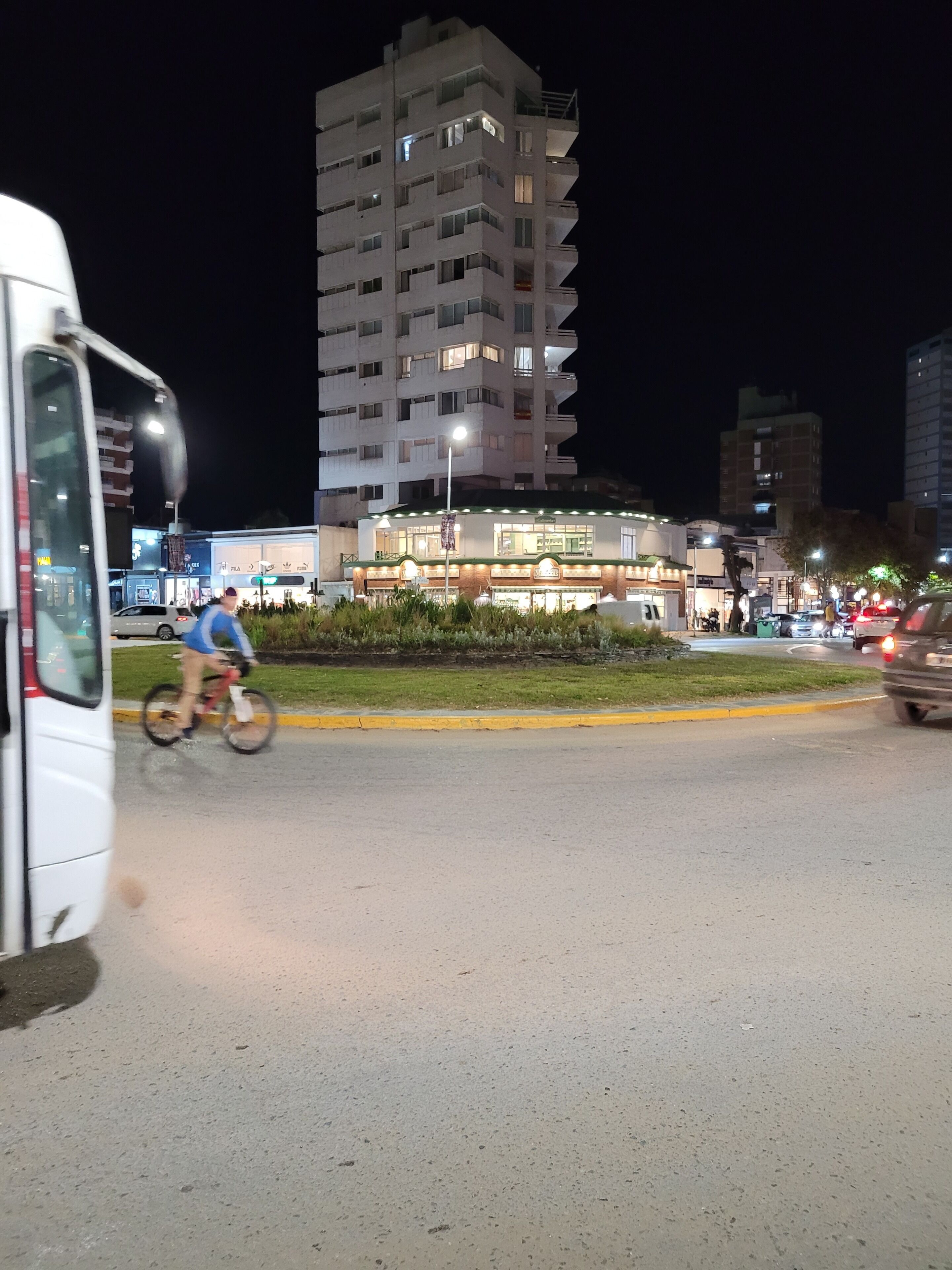 Un immeuble à plusieurs étages, doté de fenêtres illuminées et d’une entrée bien éclairée, entouré d’une zone verdoyante et bordé d’une rue animée où circulent des véhicules et un cycliste.