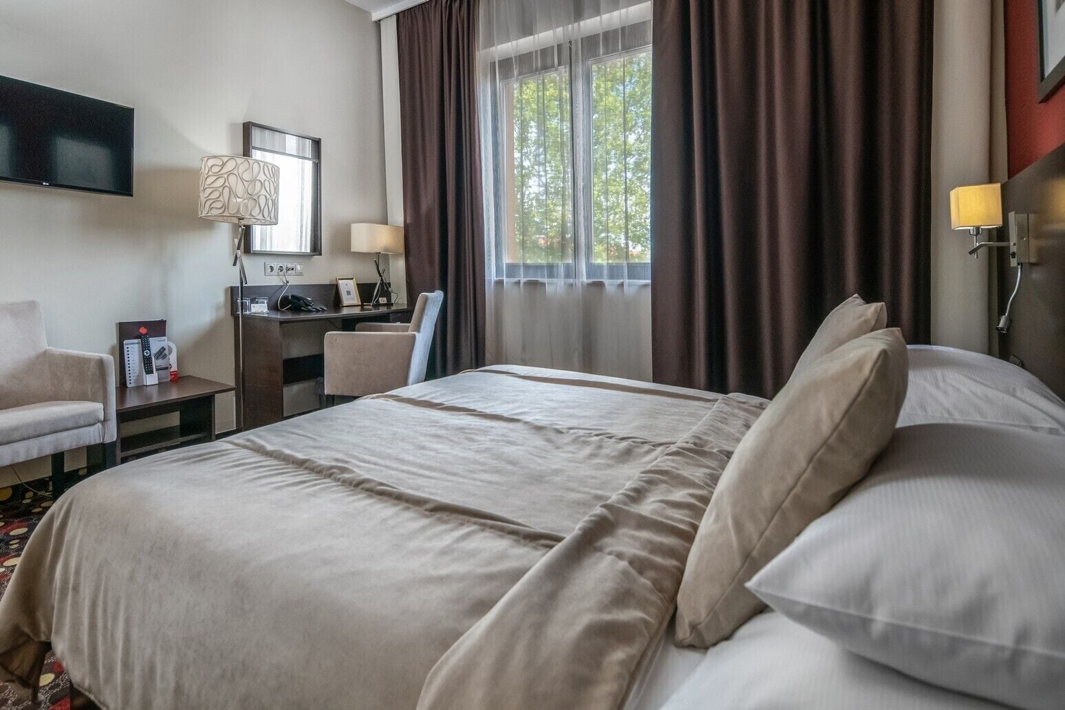 Habitación de hotel con una cama grande, un escritorio con computadora, una silla, un televisor y una ventana con cortinas.